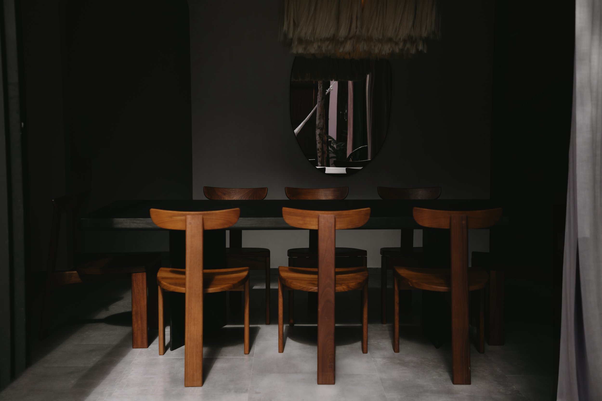 Dark dining room with a black table, six wooden chairs, a mirror on a dark wall, and minimal natural light.