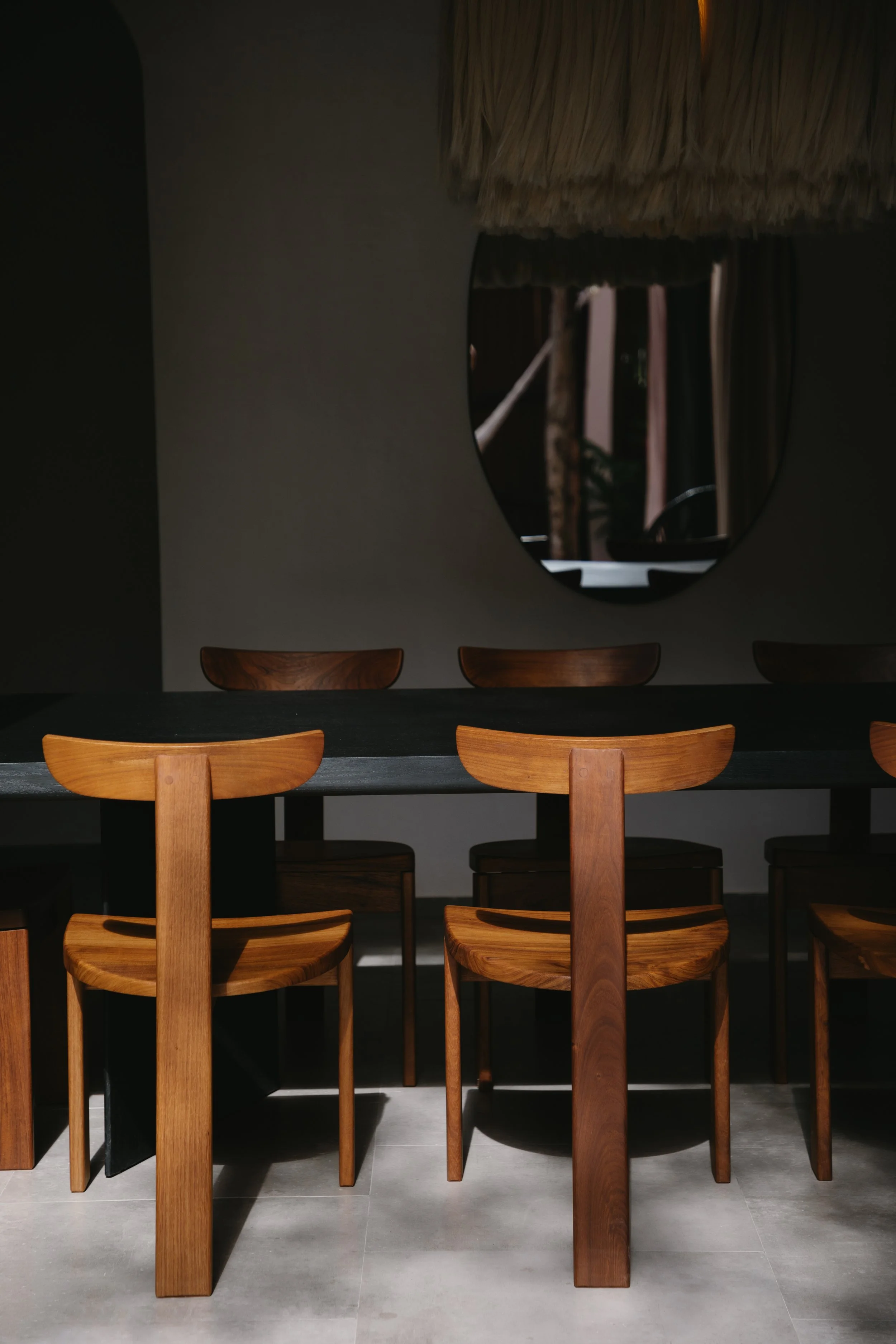 Empty dining room with wooden chairs around a dark table, shadowed interior, and a large oval mirror on the wall reflecting curtains and plants.