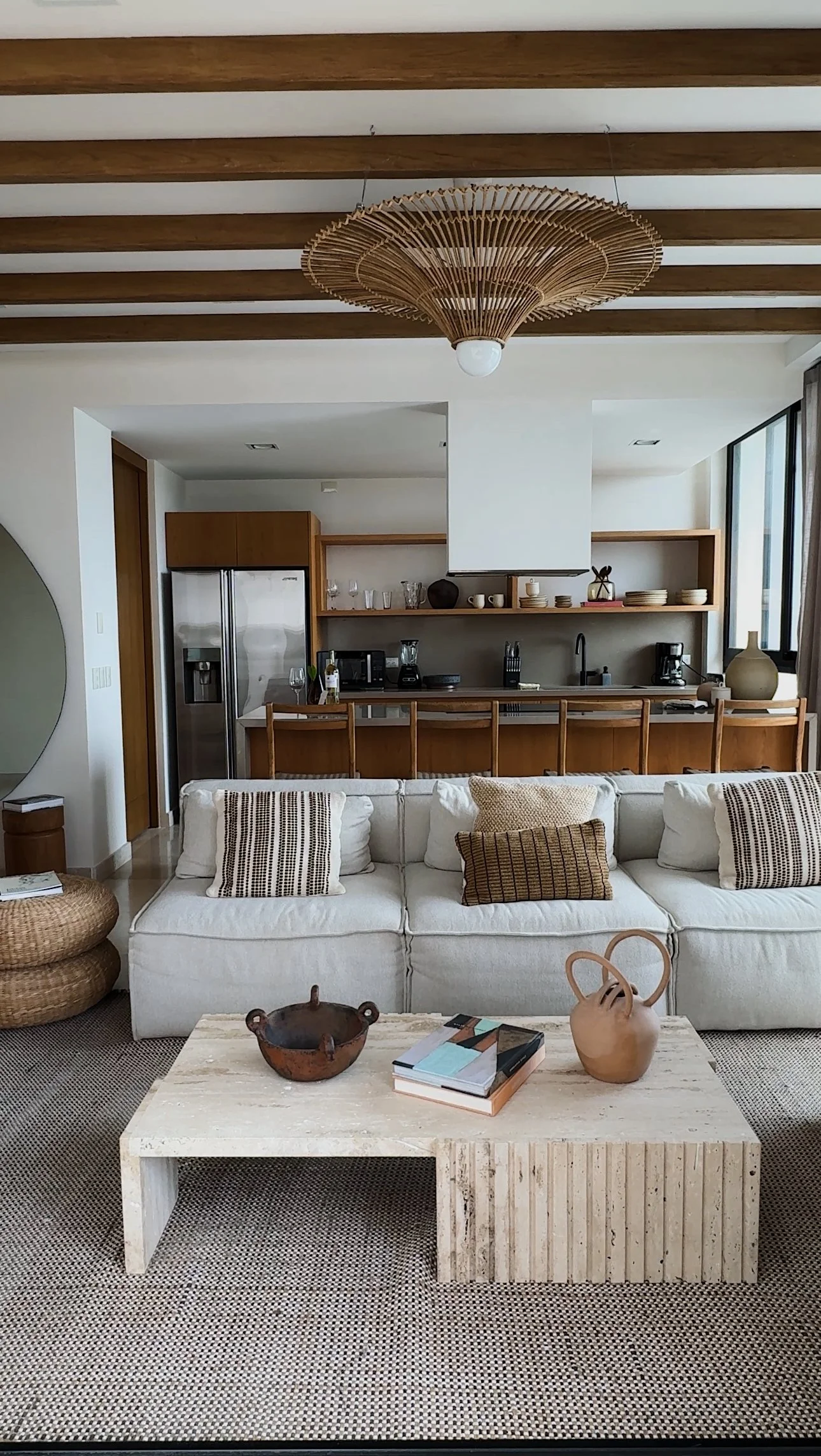 Living room with a white sofa decorated with striped pillows, a textured coffee table with books and decorative bowls, in front of an open kitchen with wooden shelves, a refrigerator, and large windows.