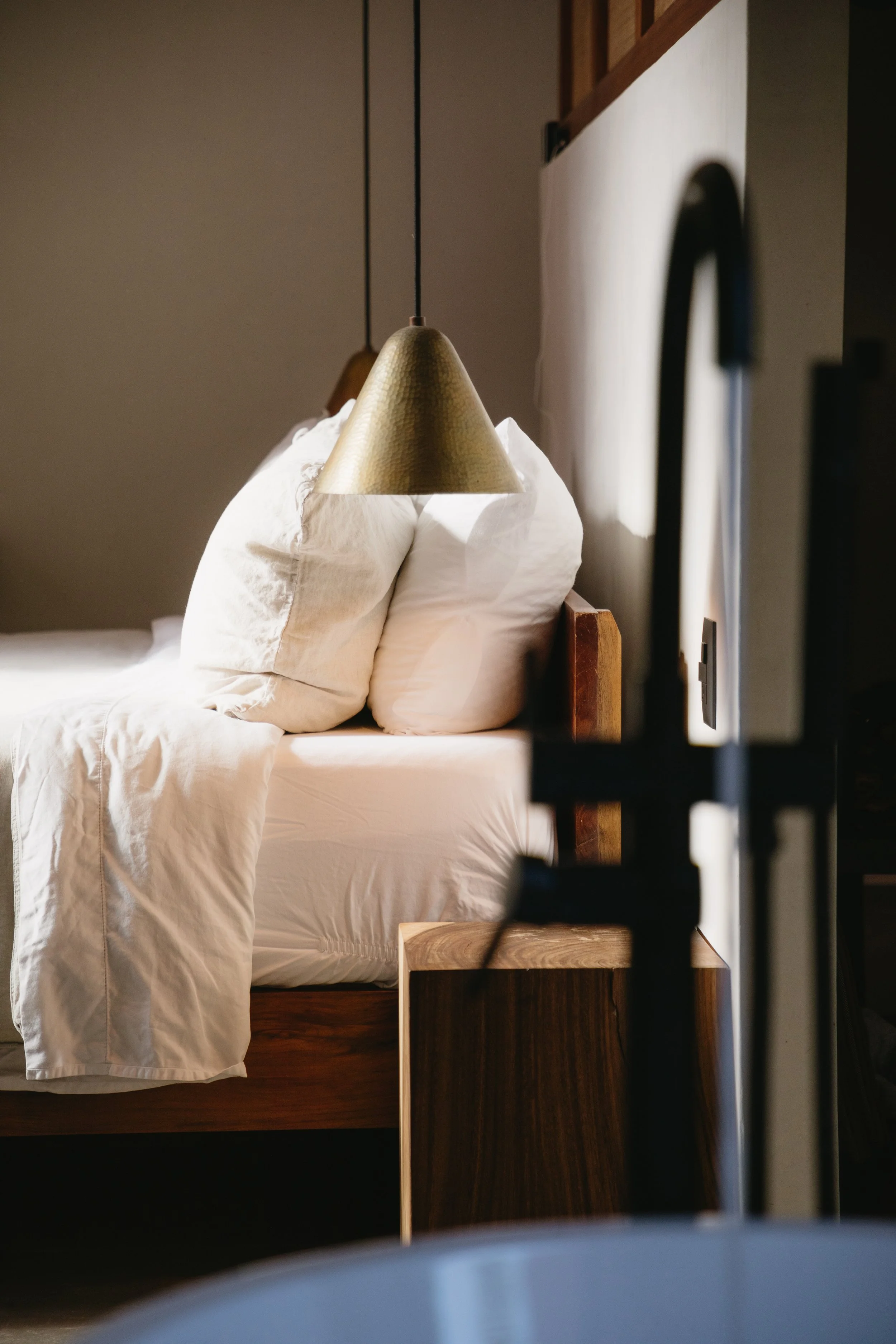 A bed with white sheets, pillows, and a beige bedspread, in a room with wooden furniture and a hanging gold-colored lamp.