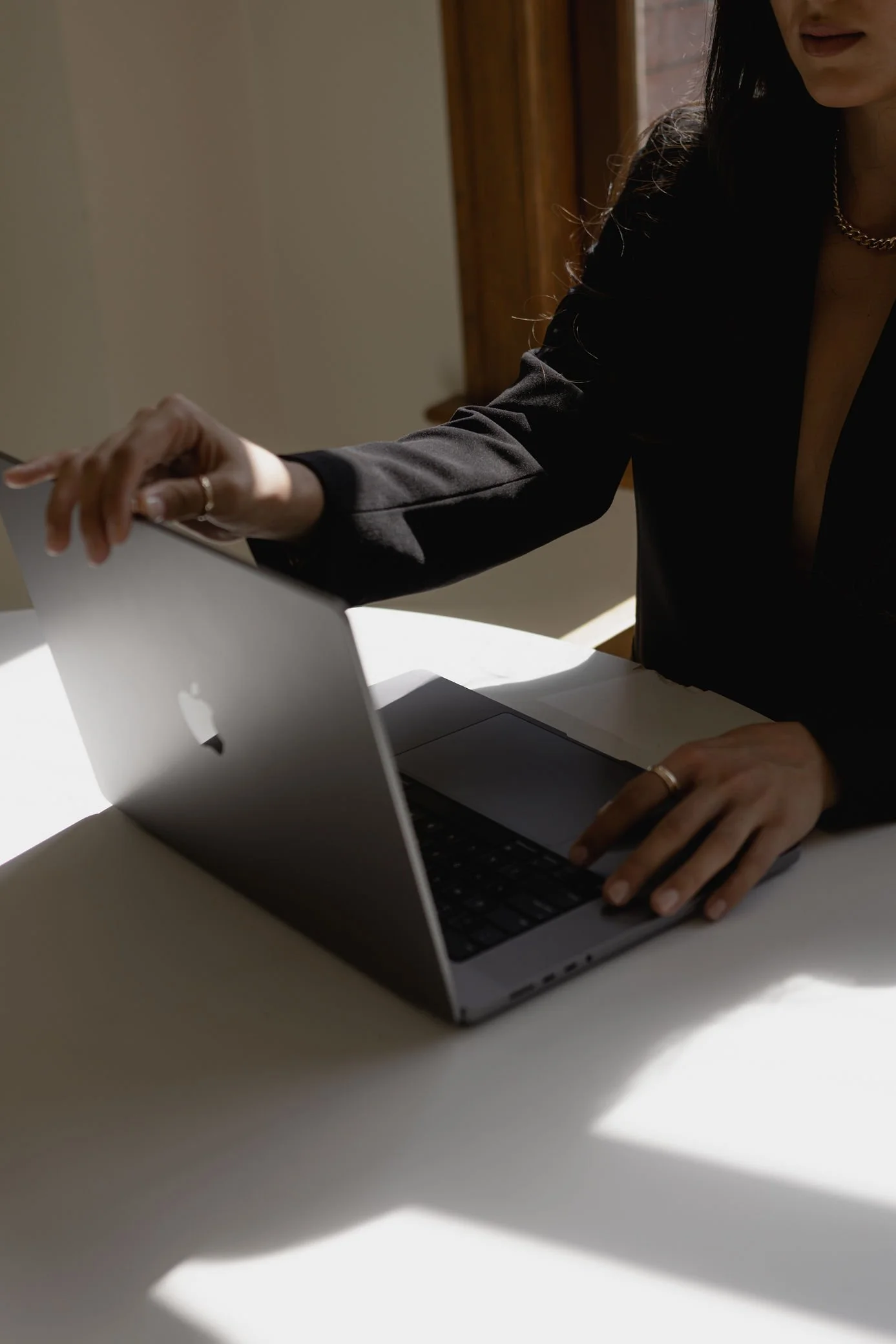 A woman with long dark hair in a black blazer working on a silver MacBook laptop at a white desk, with sunlight casting shadows on the desk.