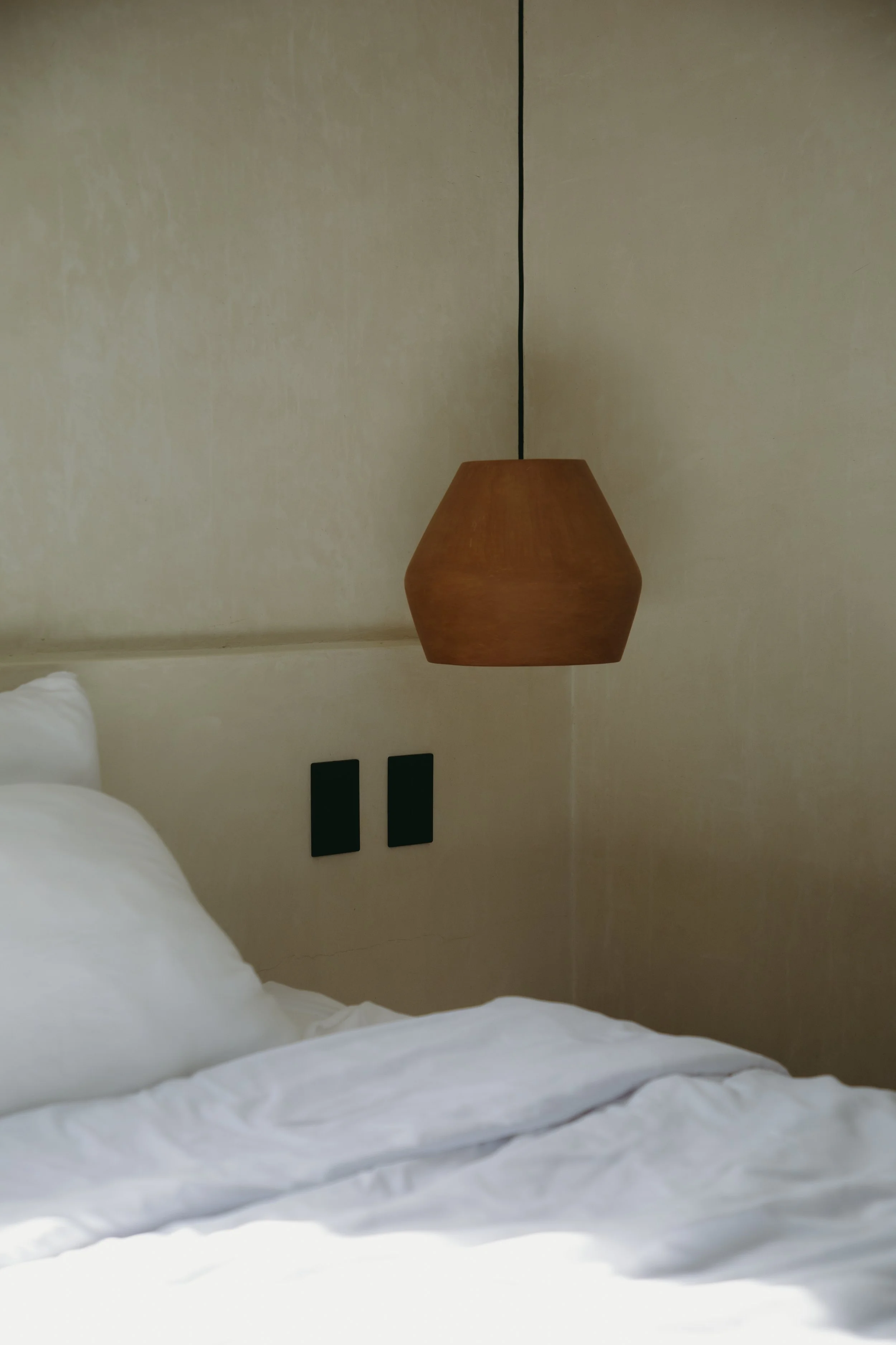 A bedroom with a hanging wooden lampshade, a bed with white bedding, two black switches on the wall.