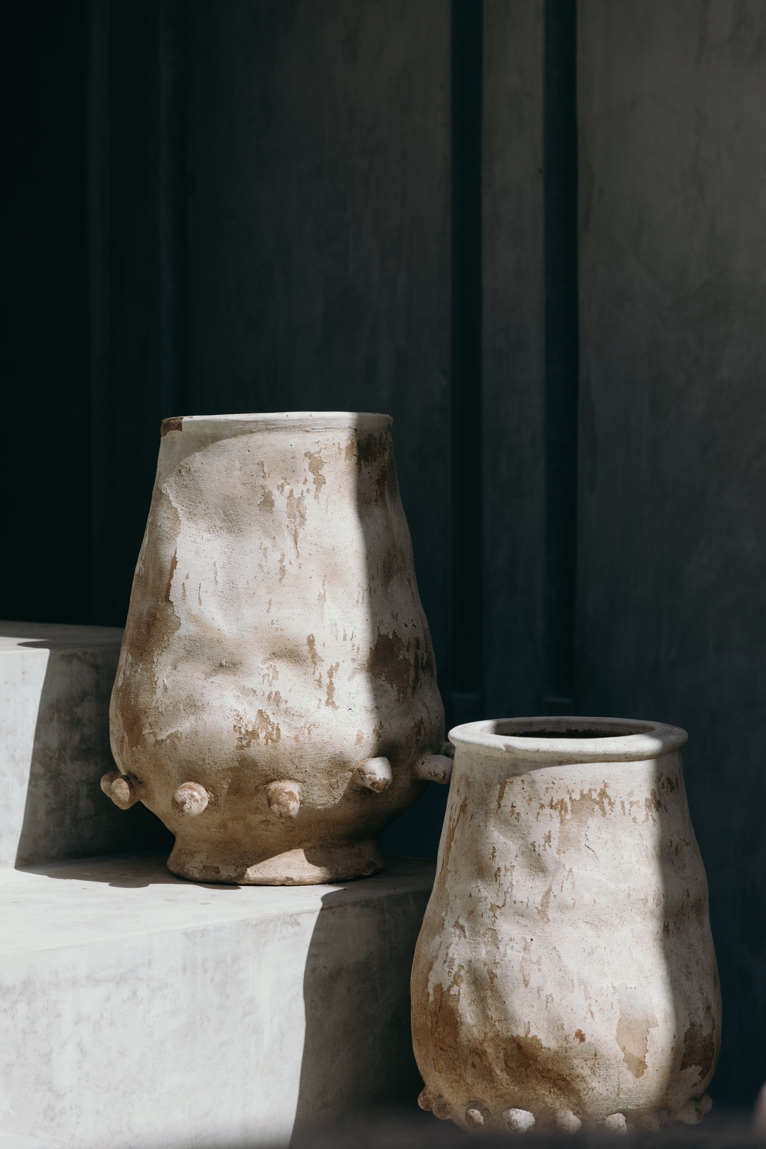 Two rustic, weathered ceramic vases with small protrusions near the base, placed on a white surface against a dark background.