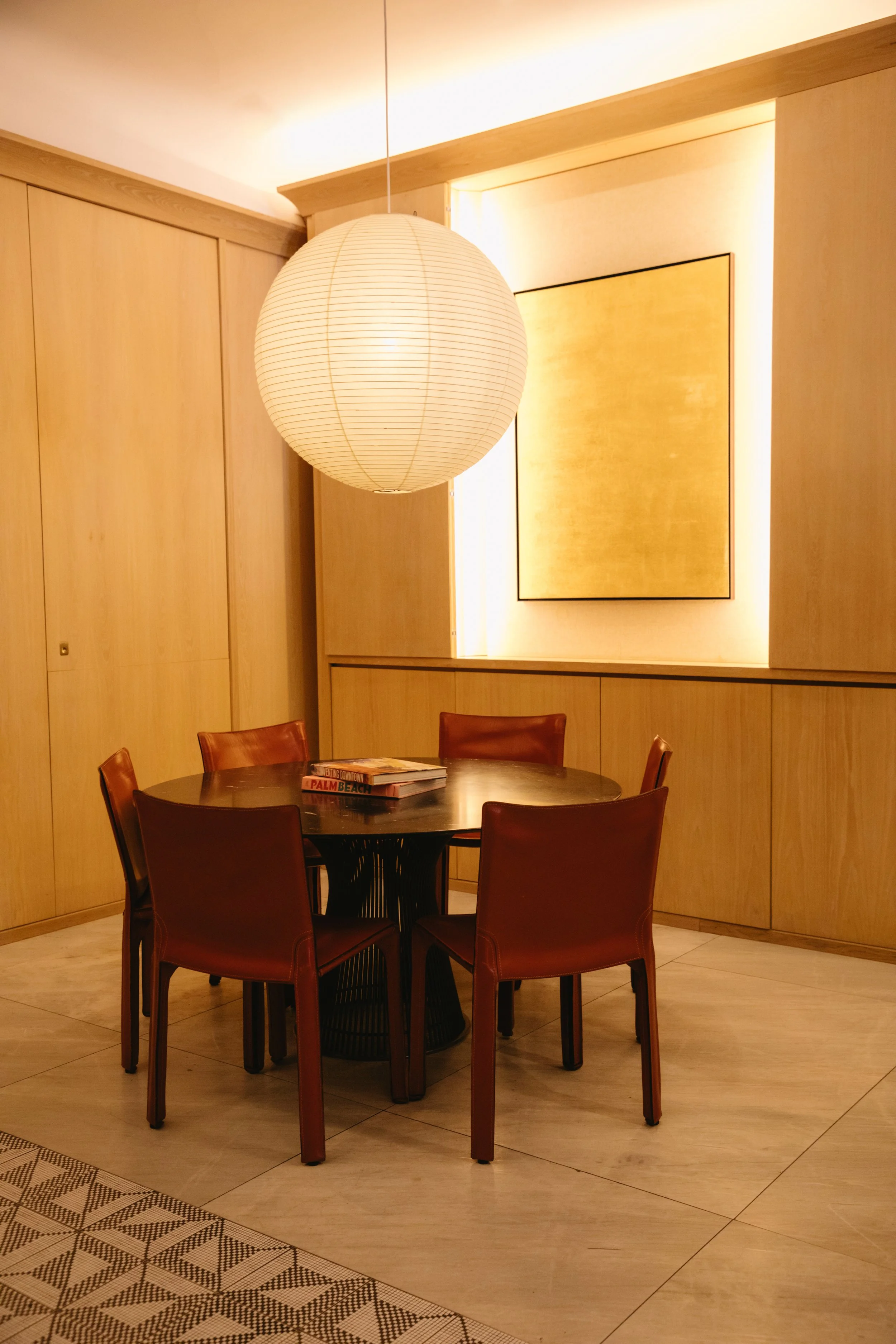 A round wooden dining table surrounded by five brown leather chairs, with a large white paper lantern hanging overhead, set in a softly lit room with wooden paneling and a yellow artwork on the wall.