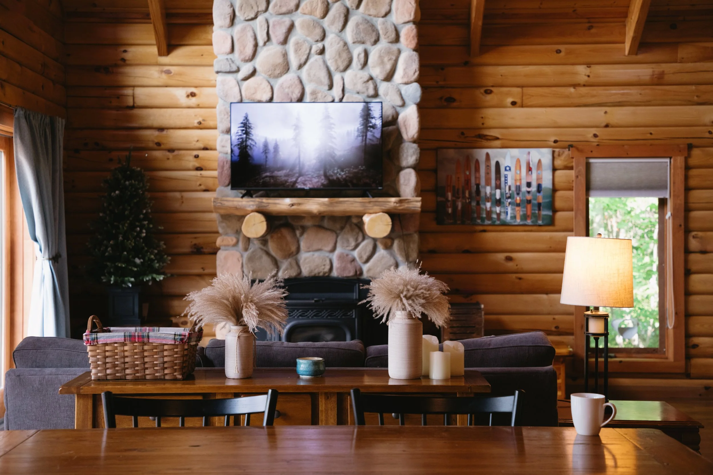 Living room with wooden walls, a stone fireplace with a television on top, a small Christmas tree to the left, and a window with a view of greenery. There are pillows, candles, a lamp, and a coffee mug visible.