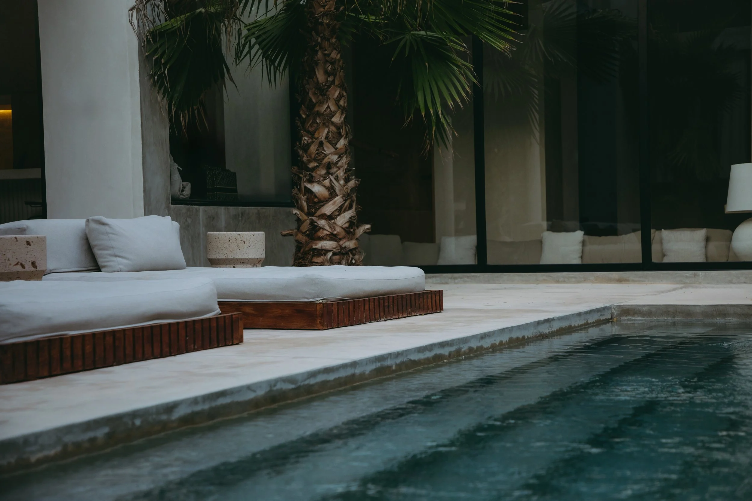 A modern outdoor pool area with white cushioned lounge beds, a palm tree, large glass windows, and outdoor seating visible through the windows.