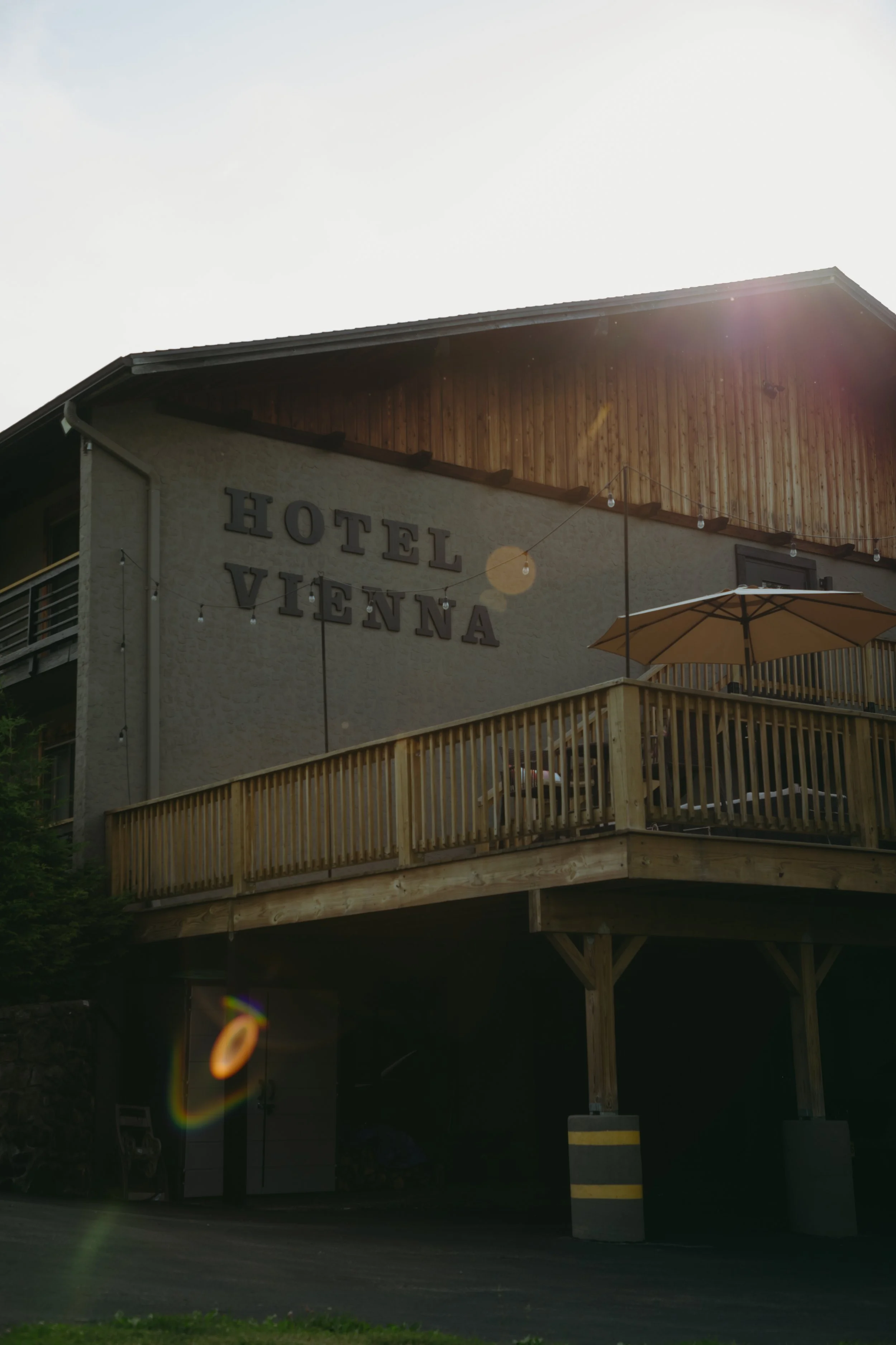 Exterior of a hotel building with a wooden balcony, string lights, and a sunburst effect, with the sign 'Hotel Vienna' on the wall.