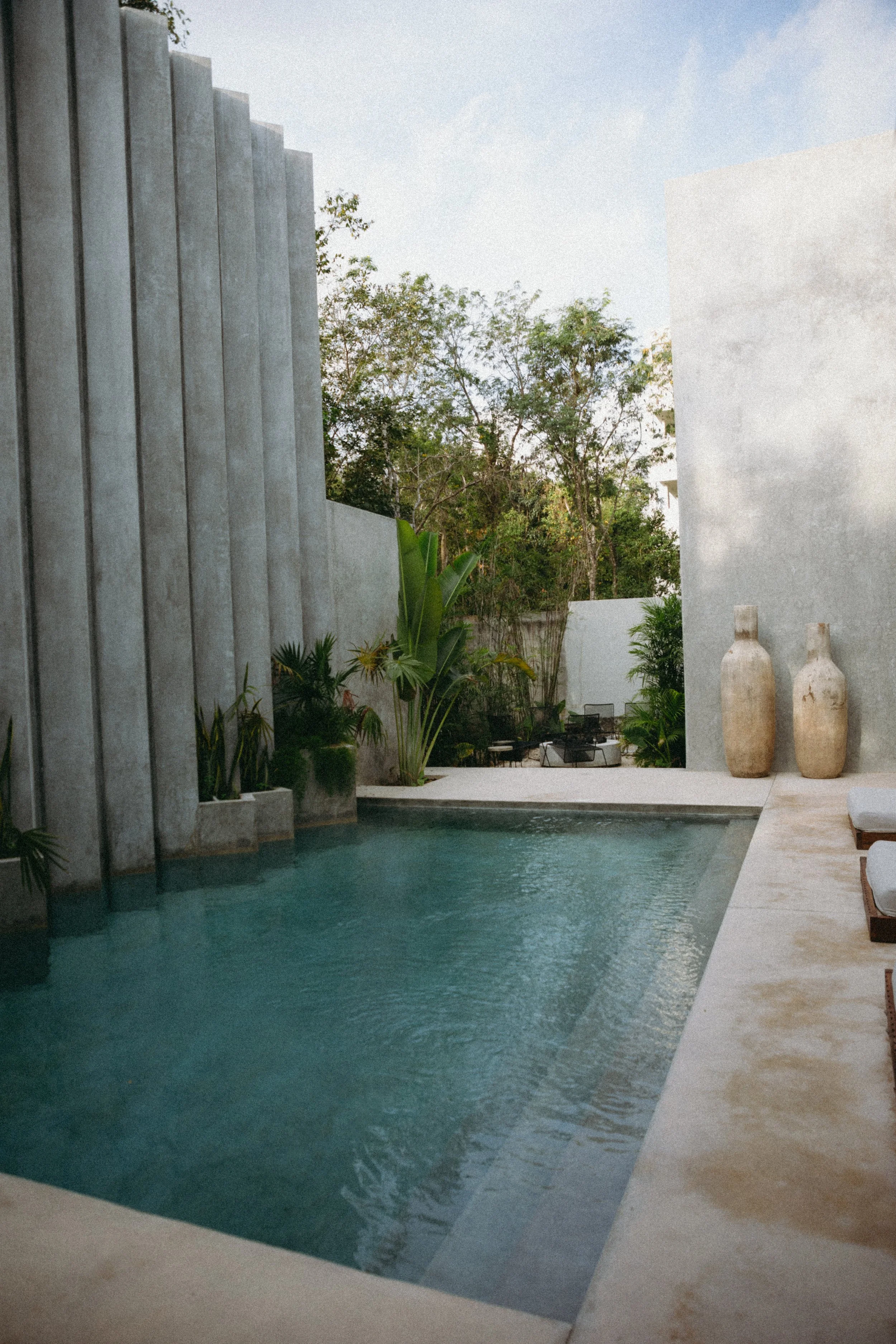 A modern outdoor pool area with concrete walls, greenery, and decorative vases, under a partly cloudy sky.