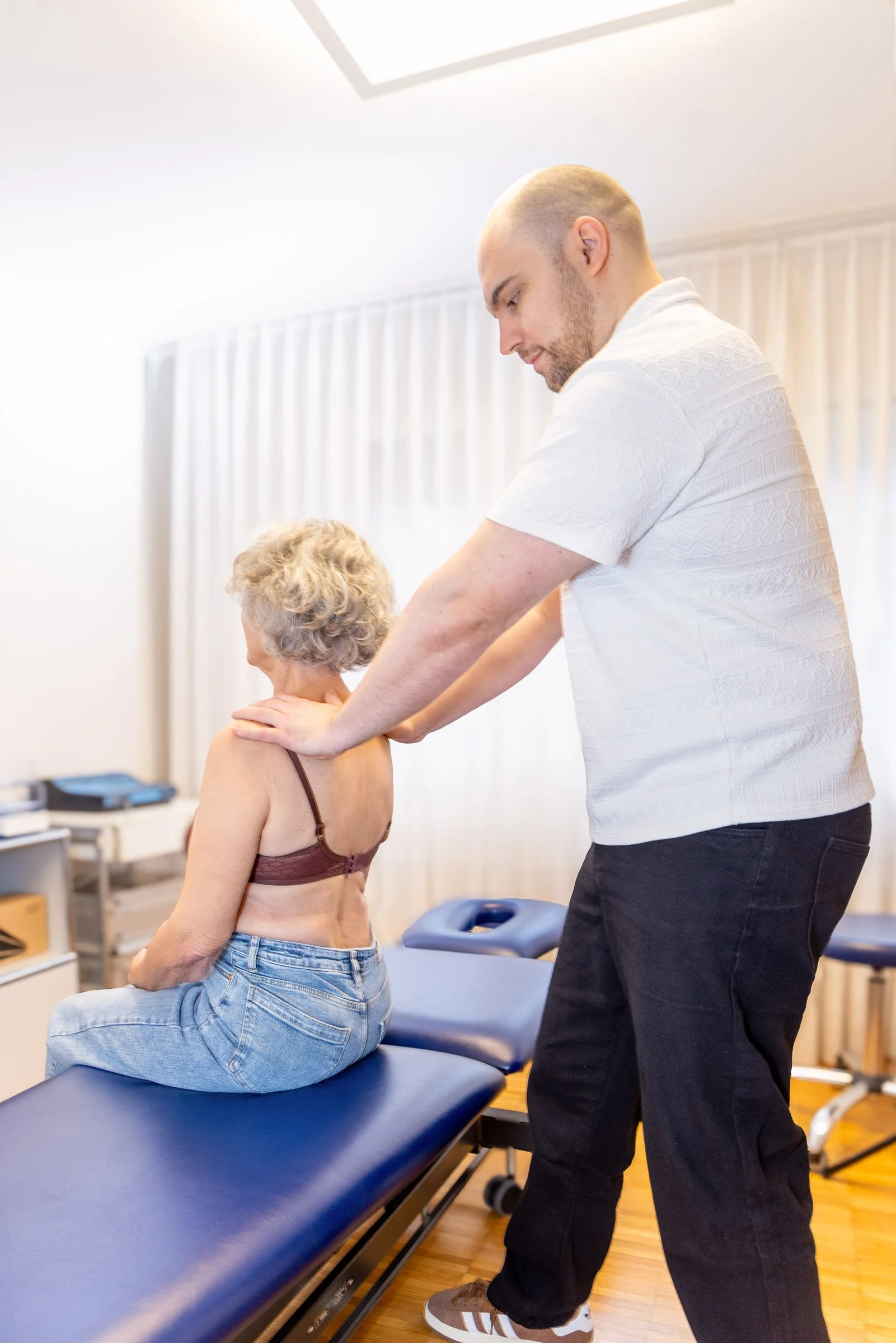 Ein Mann massiert eine ältere Frau, die auf einem Untersuchungsliege sitzt, im Behandlungsraum.