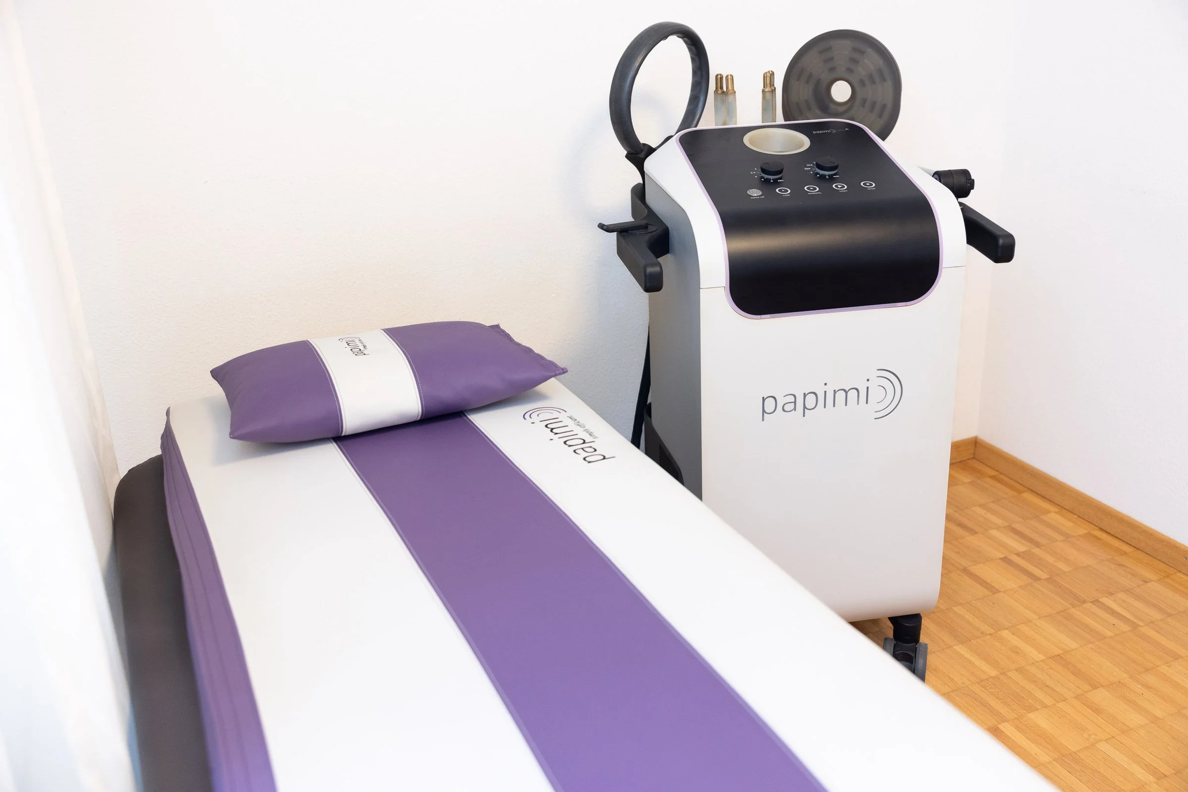 Behandlungsraum mit medizinischem Gerät und Behandlungsbett, das mit einem lila und weißen Bezug bedeckt ist.