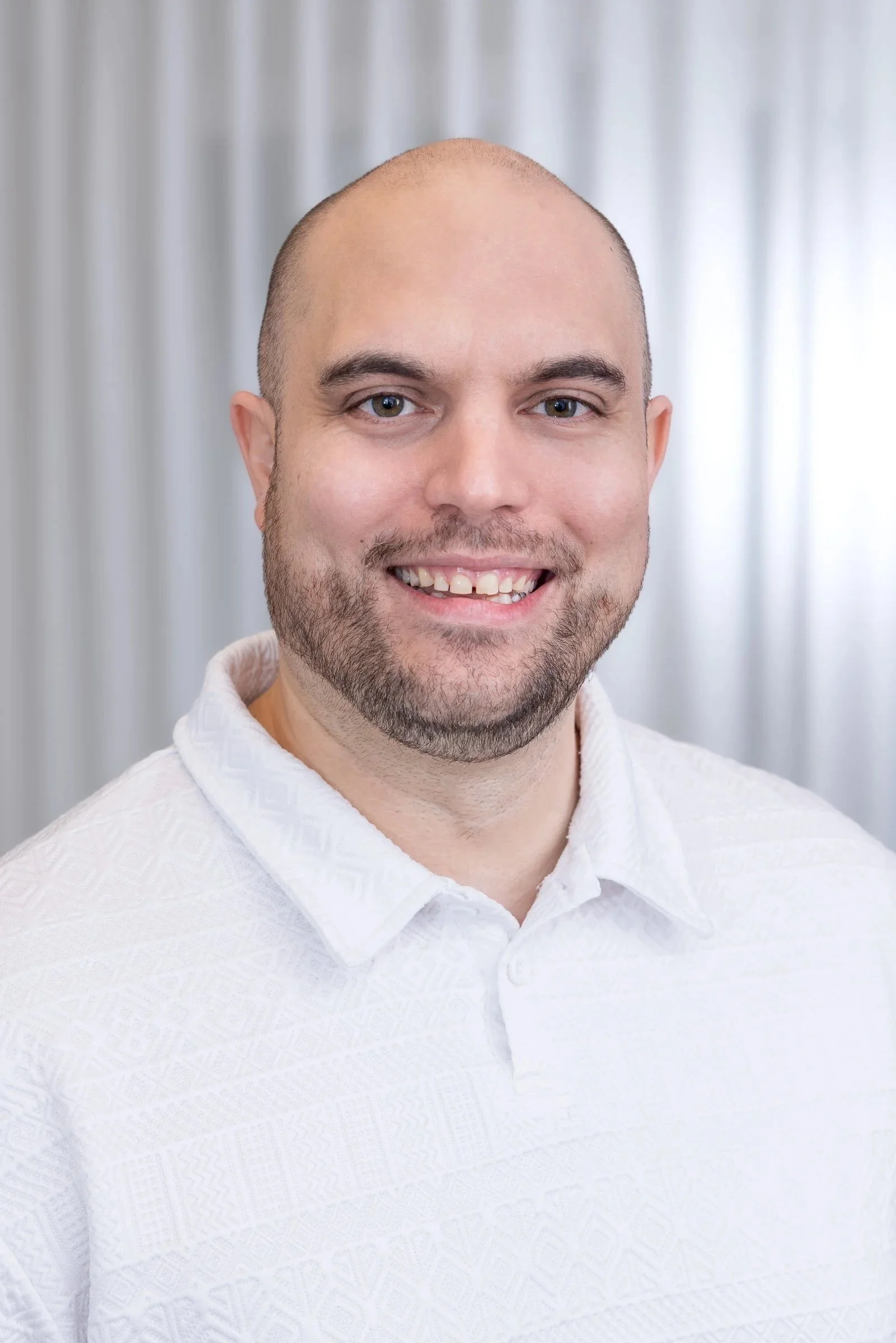 Nahaufnahme eines lächelnden Mannes mit kurzen dunklen Haaren, Bart und blauen Augen, trägt ein weißes Hemd, vor einem metallischen Hintergrund.