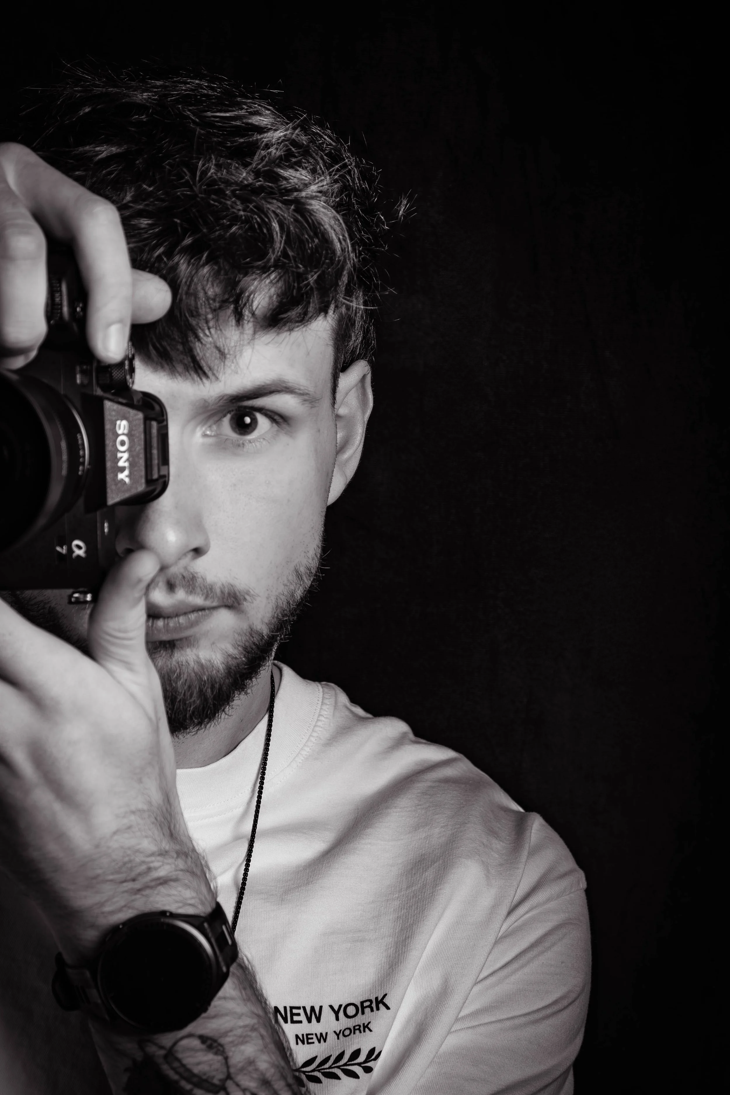 Jeune homme avec une barbe et des cheveux bouclés, tenant un appareil photo Sony, en train de prendre un autoportrait en noir et blanc.