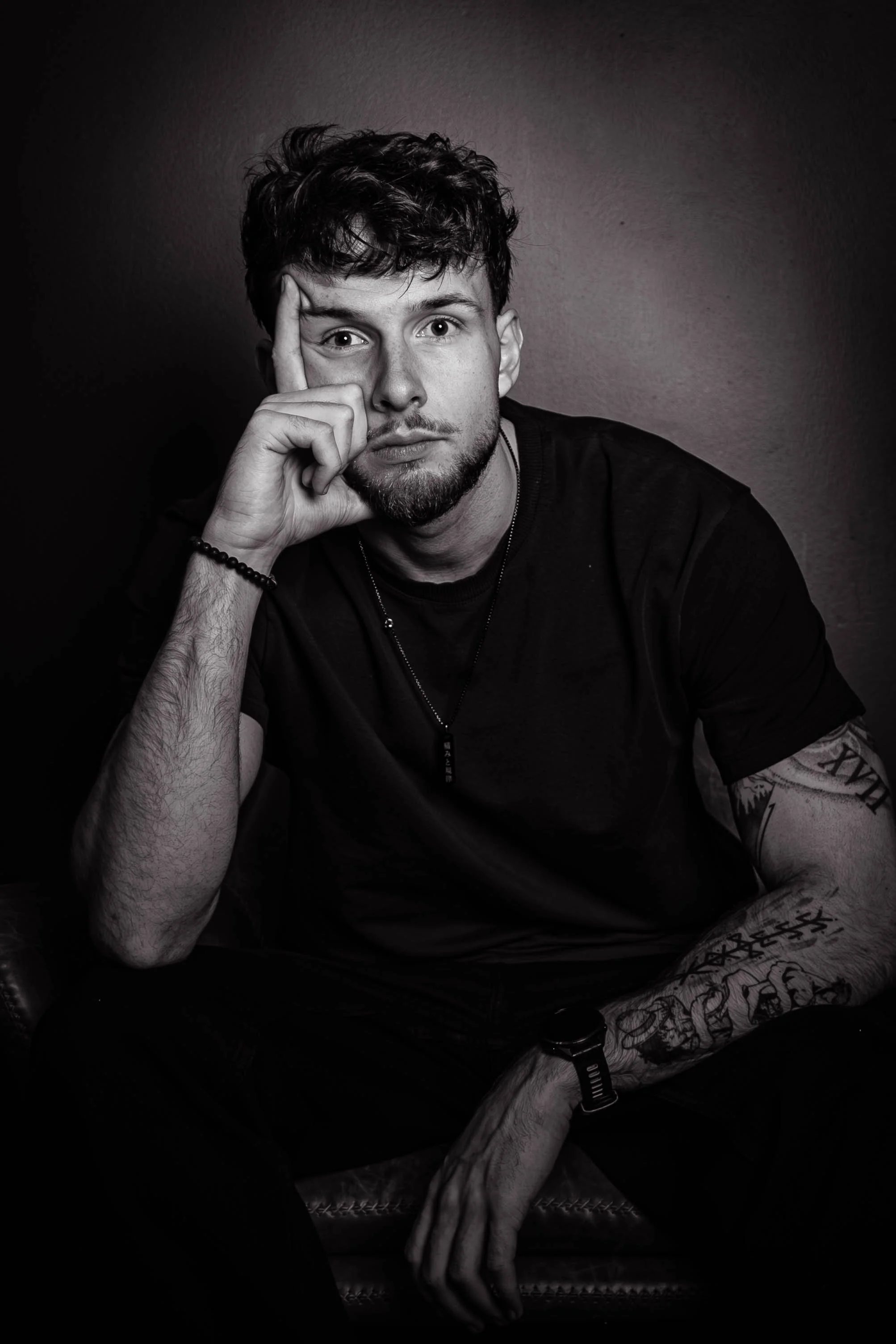 Un jeune homme aux cheveux bouclés assis, portant un t-shirt noir, avec des tatouages sur le bras gauche et un bracelet, posant avec la main sur son visage, dans un style monochrome.