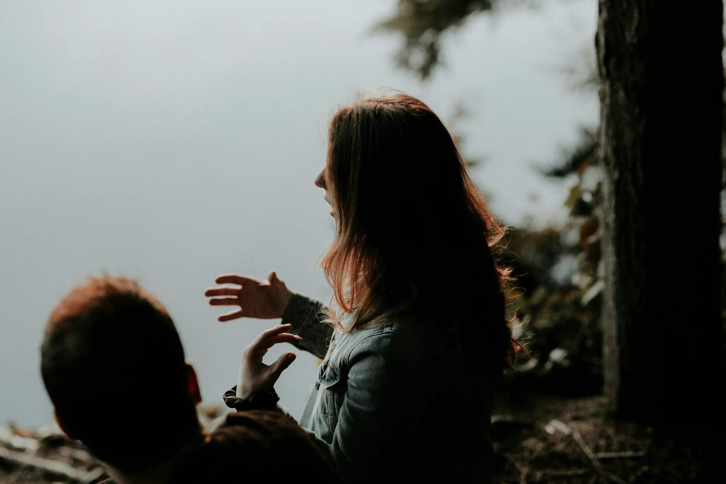 A woman with wavy hair sitting outdoors near a tree, gesturing with her right hand, with a person in the foreground whose back is facing the camera, during daytime.