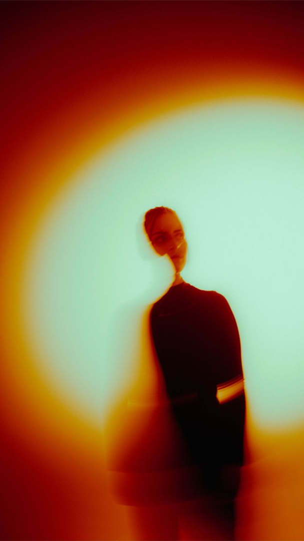 Person standing behind a circular light or projection, creating a glowing effect around them with a distorted face due to the light.