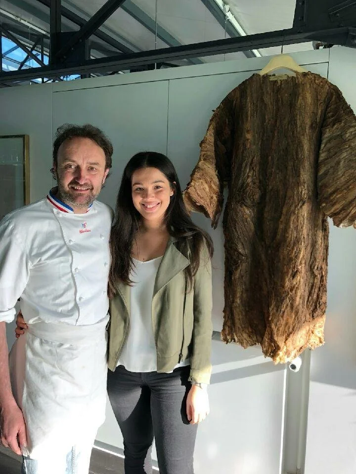 Deux personnes posent à côté d'une grande pièce de cuir ou de foin suspendue dans un espace d'exposition ou un musée.