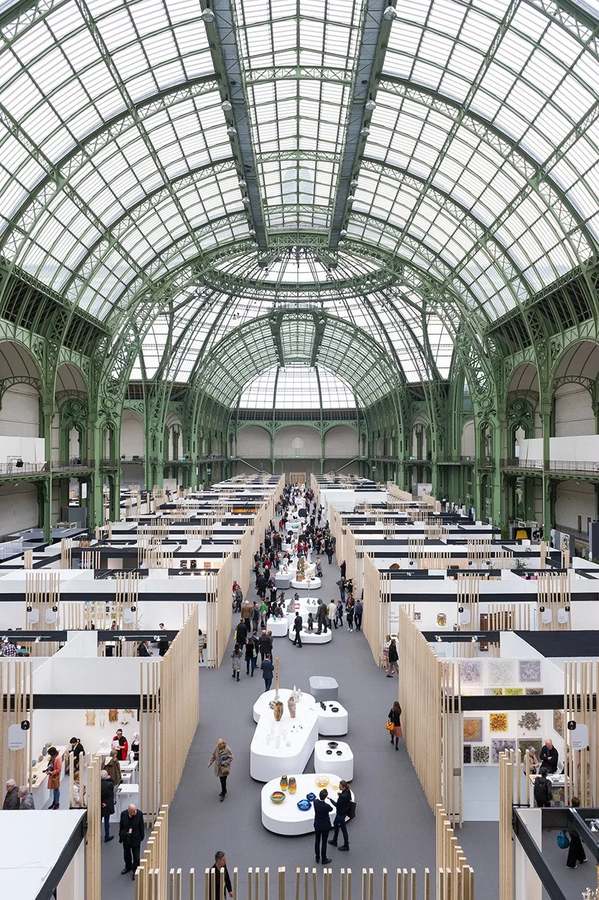 Grand marché ou exposition dans un bâtiment avec une grande verrière en acier vert, avec des stands et des visiteurs.