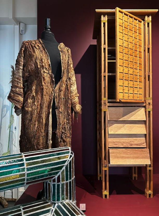 Vêtement en fourrure ou en textile imitant la fourrure, exposé sur un mannequin, à côté d'une table en mosaïque et d'une grande armoire en bois avec des portes ajarées, dans une pièce d'exposition.