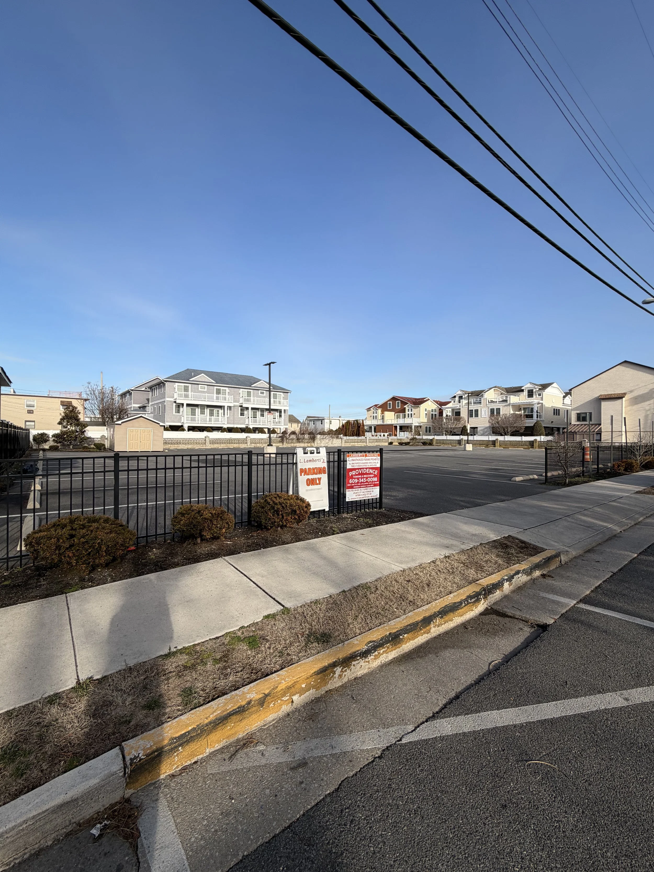 Empty parking lot with signs, small bushes, sidewalk, and residential houses in the background under a clear blue sky.