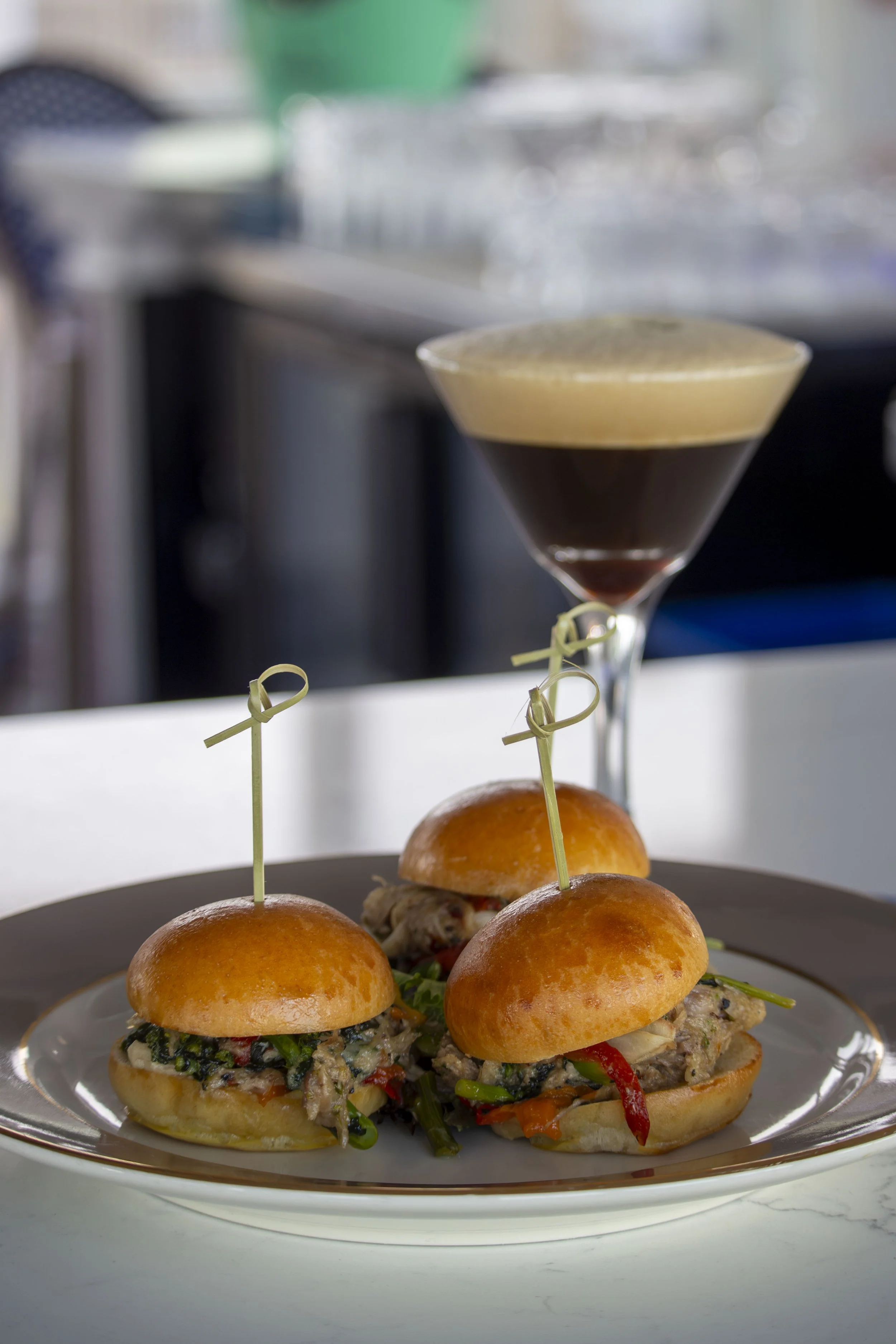 Three sliders with meat patty, vegetables, and buns on a silver plate, with a blurred background of a kitchen and a espresso martini in a martini glass sitting on a bar top