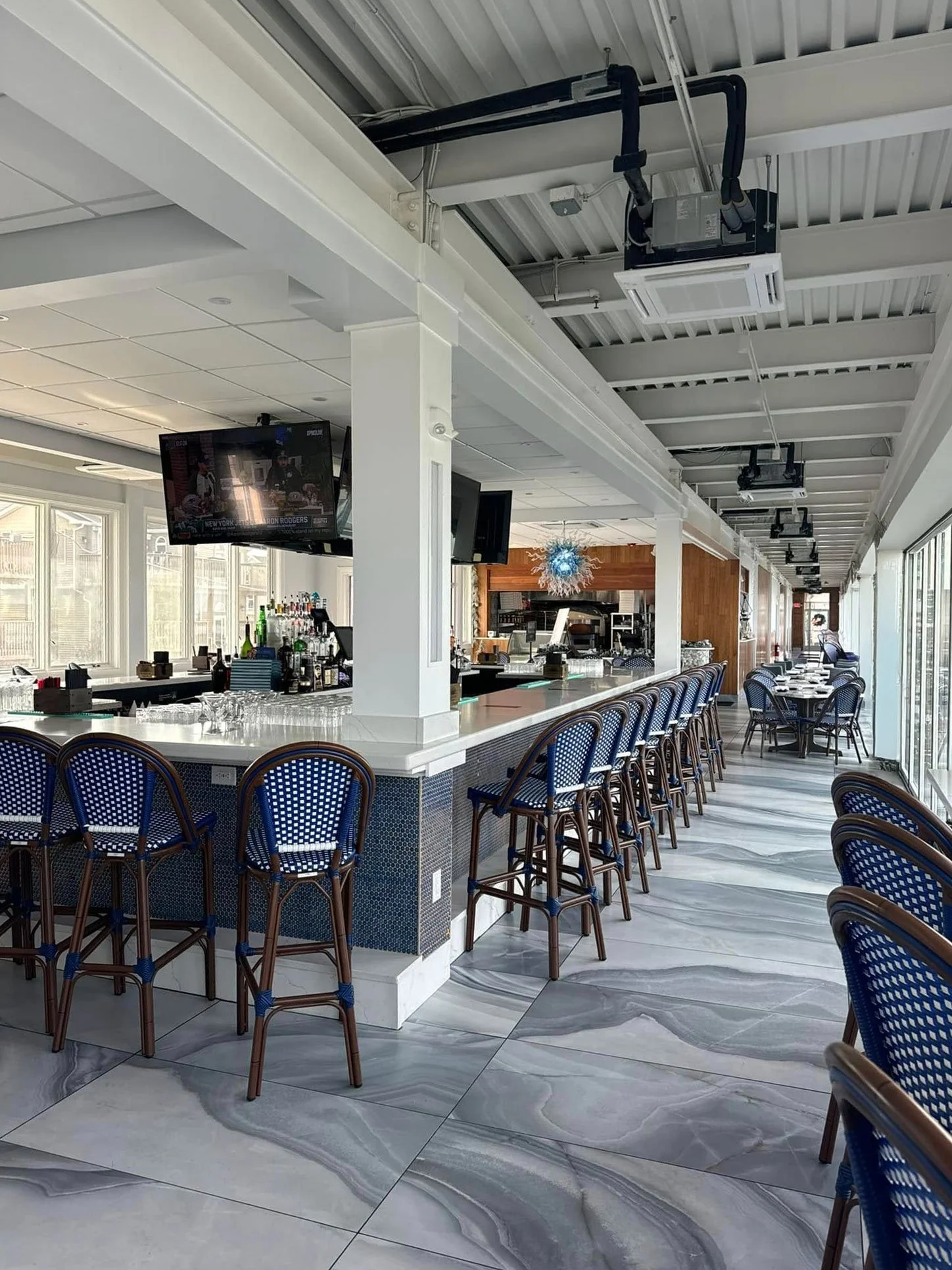 A modern indoor restaurant with a bar counter, blue and brown chairs, large windows, and a marble floor.
