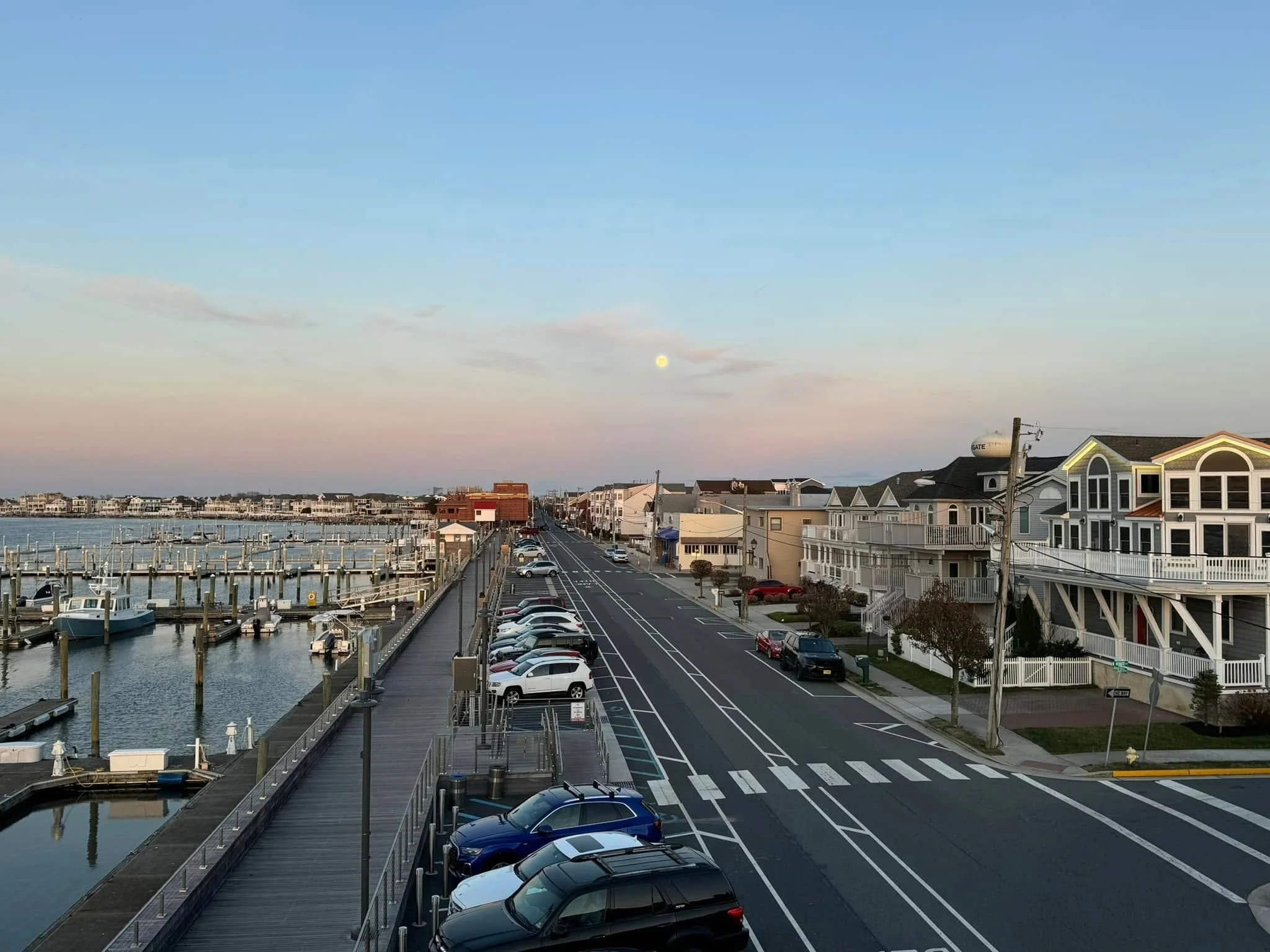 View of a marina with boats docked along piers on the left, a parking lot filled with cars in the foreground, a quiet street with residential houses on the right, and a pastel-colored sky with a visible moon during sunset.
