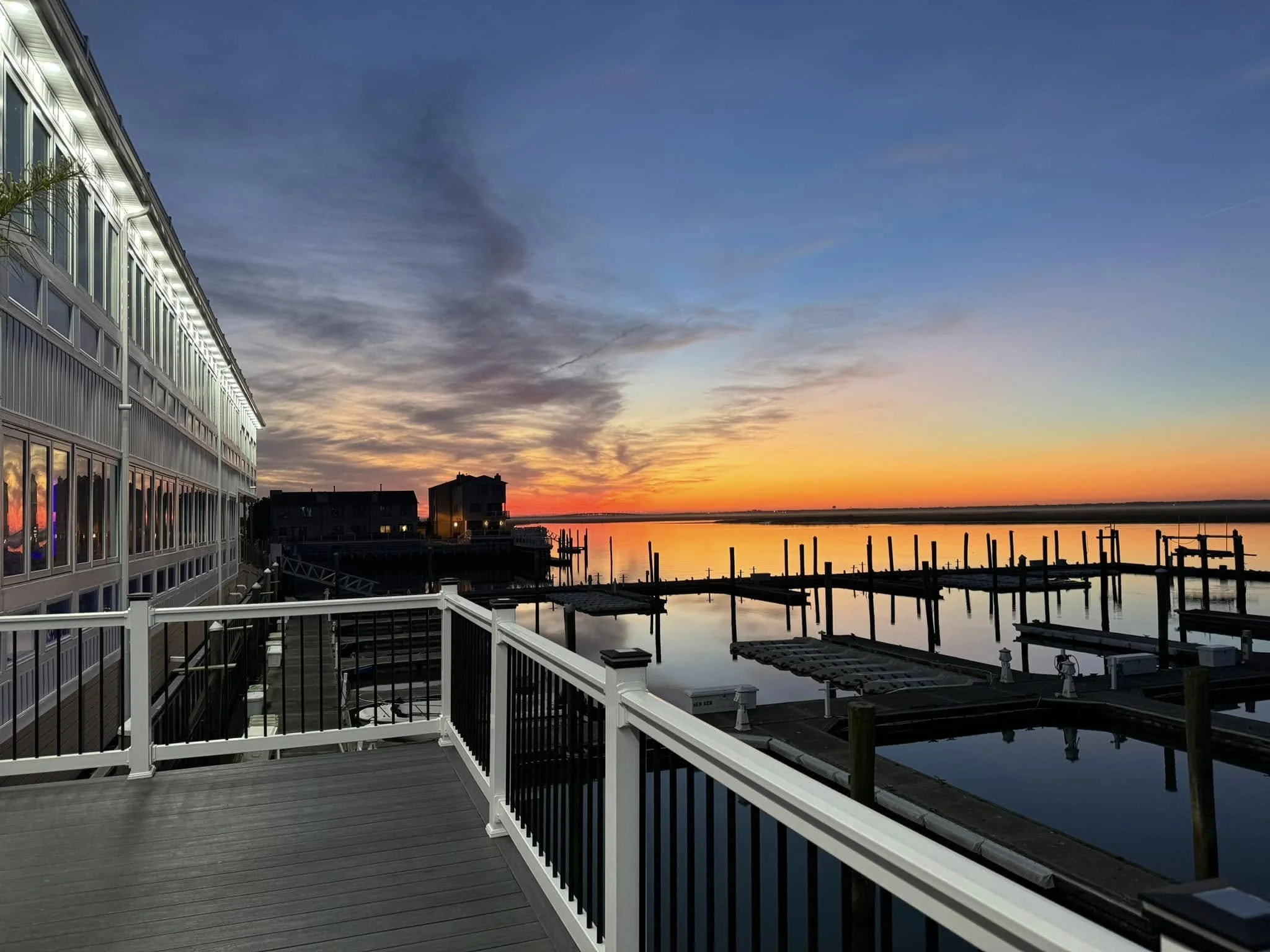 Sunset over a marina with calm water, wooden docks, and boats, seen from a balcony with a white railing, adjacent to a multi-story building with illuminated windows.