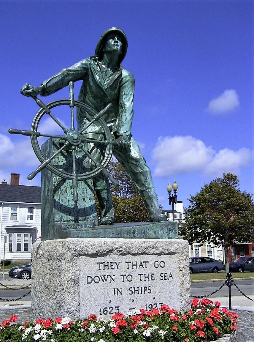 Bronze statue of a sailor steering a ship's wheel, mounted on a stone base with an inscription, surrounded by flowers, in an outdoor setting with buildings and trees in the background.