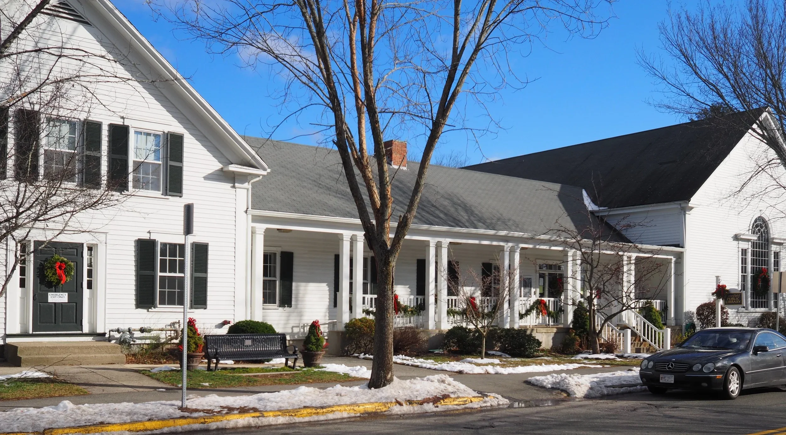 White residential building decorated with holiday wreaths and garlands, with leafless trees in front, a bench, and a sports car parked on the street, under a partly cloudy sky.