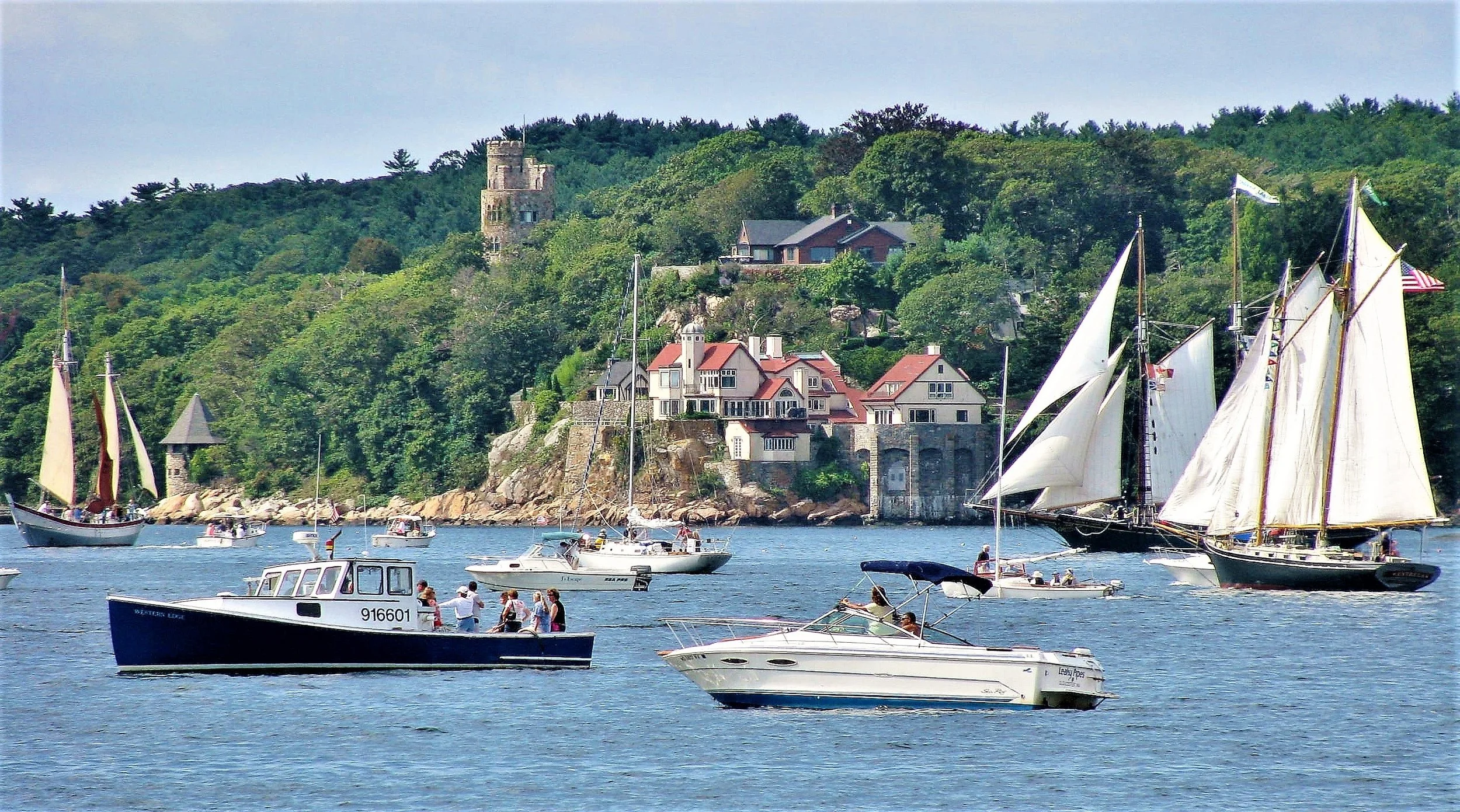 Boats sailing on a body of water near a hillside with houses and a stone tower surrounded by trees.