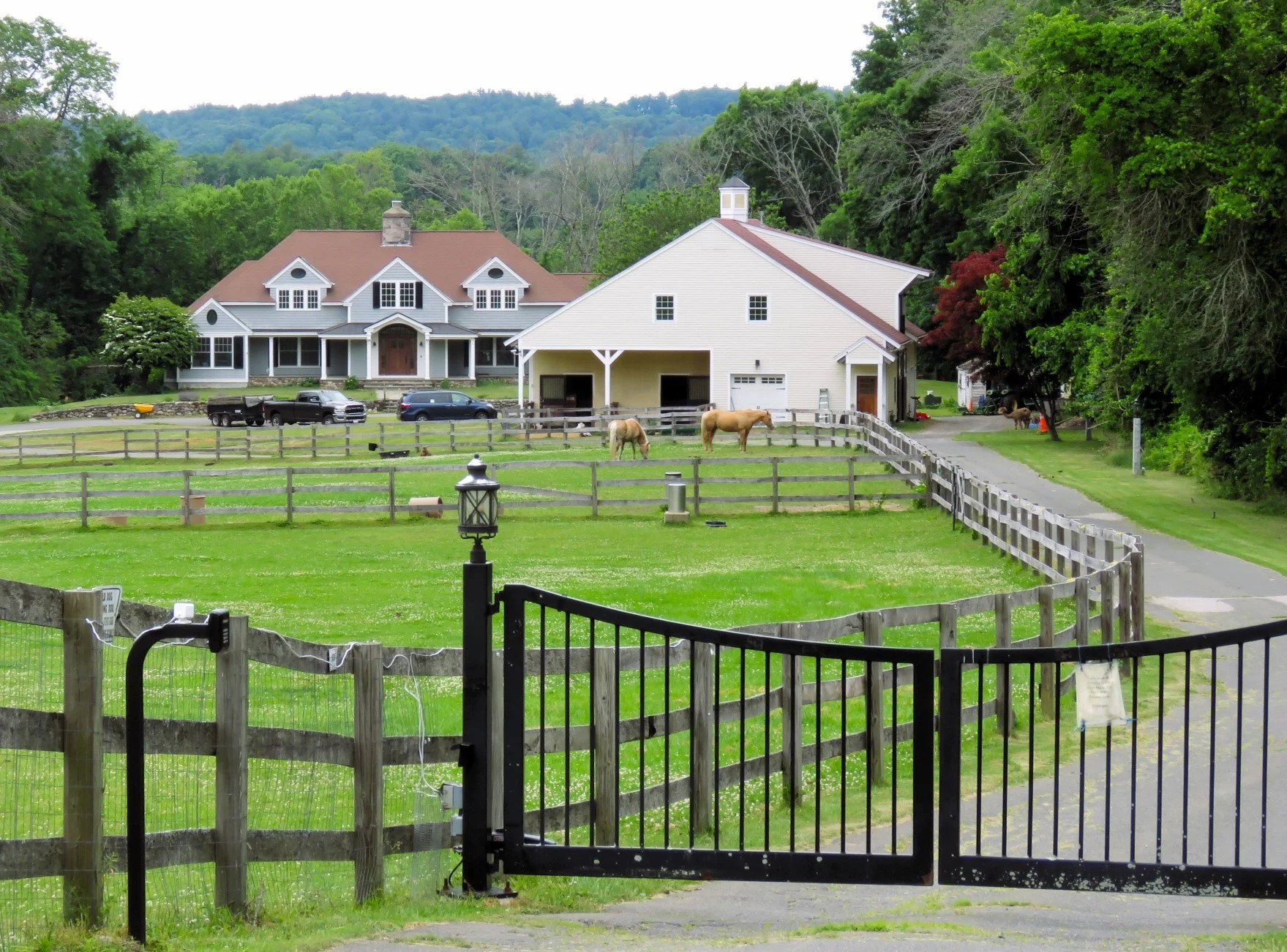 A large house with a brown roof and white exterior, situated behind a fenced yard with green grass on a farm or rural property. In the yard, there are two horses grazing and several vehicles parked nearby. The background includes dense green trees and a mountain or hill in the distance.