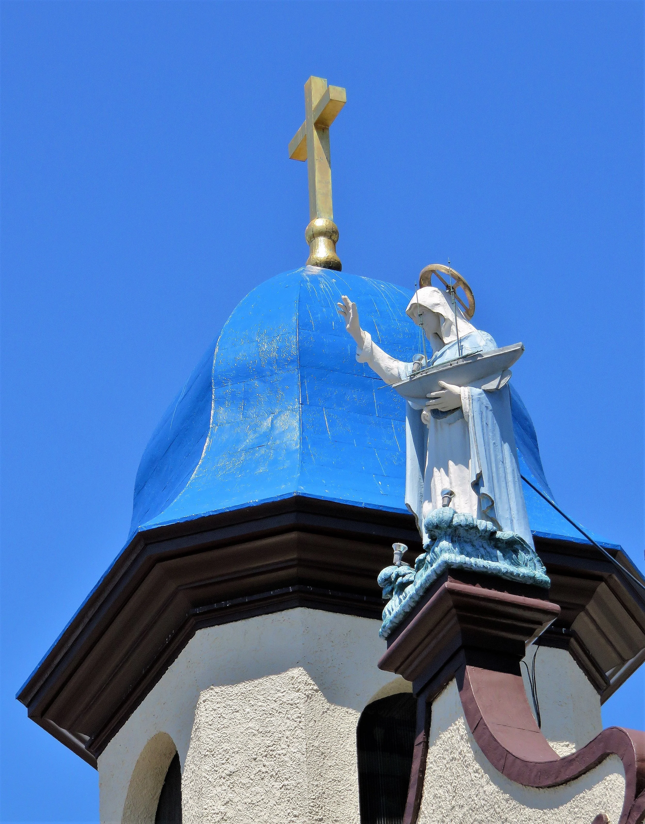 Statue of a religious figure holding a book with a cross and a wheel above, atop a building with a blue dome and a decorative trim, against a clear blue sky.