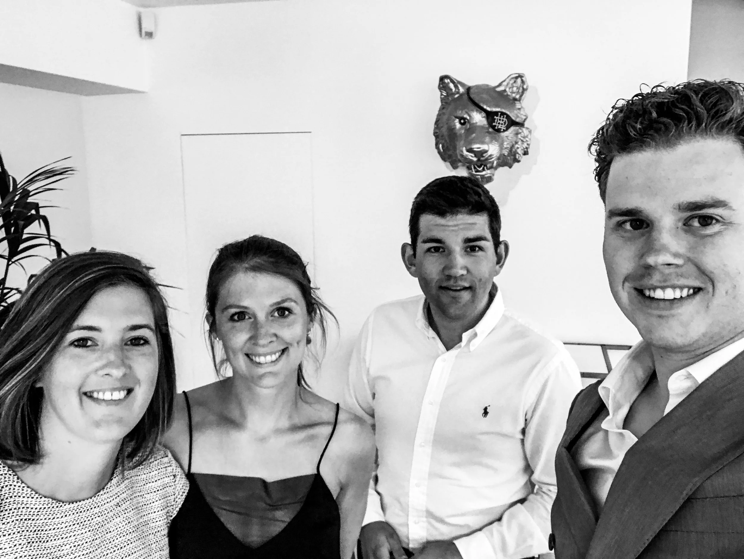 Four young adults smiling for a photo in a room with a wolf head wall decoration with an eye patch.