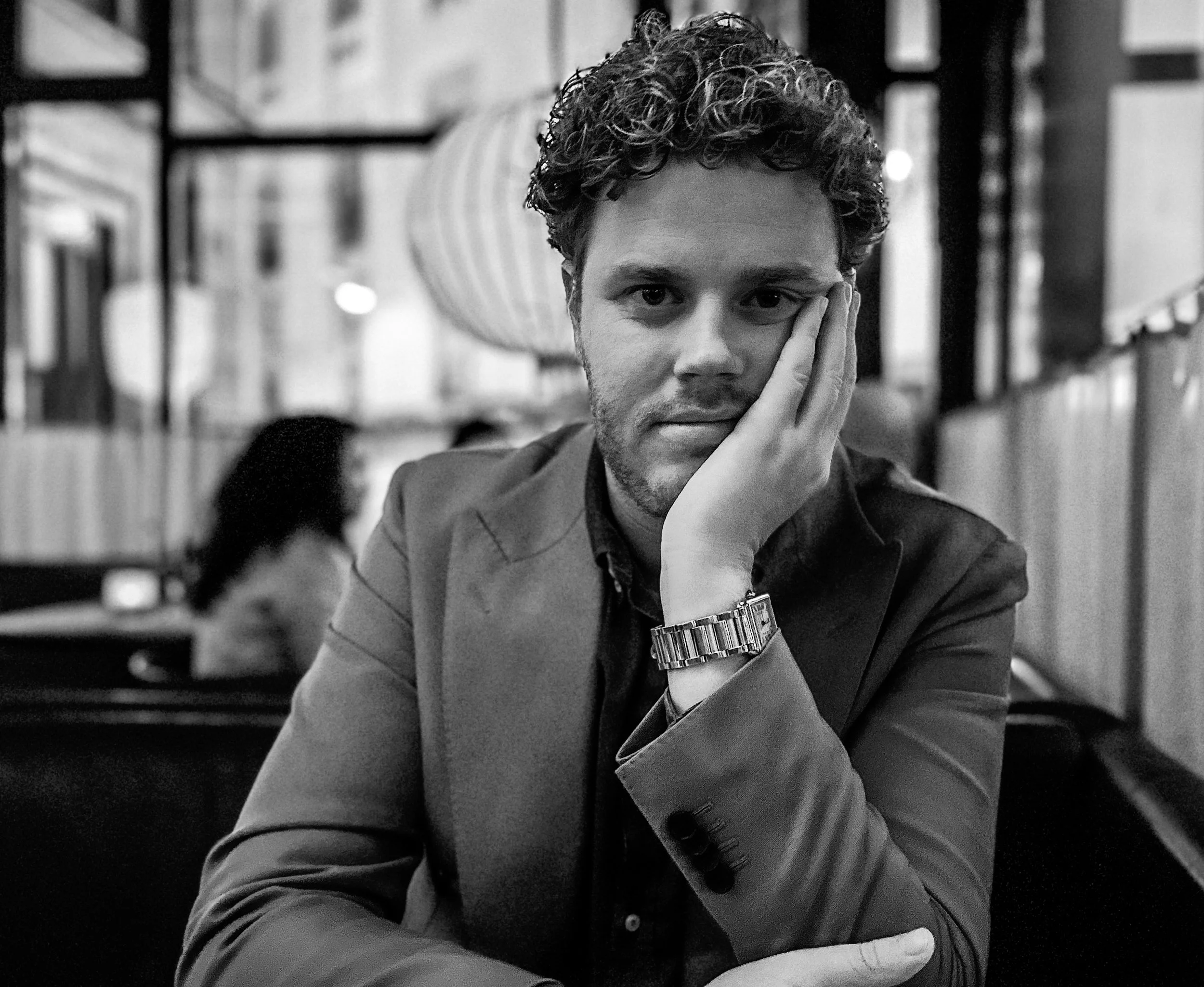 A black-and-white photo of a man with curly hair and a beard, sitting at a restaurant table, resting his head on his hand, looking directly at the camera.