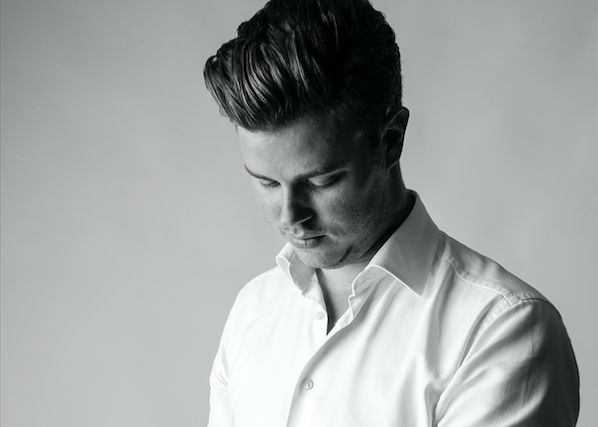 Black and white photo of a young man with styled hair wearing a white dress shirt, looking downward against a plain background.