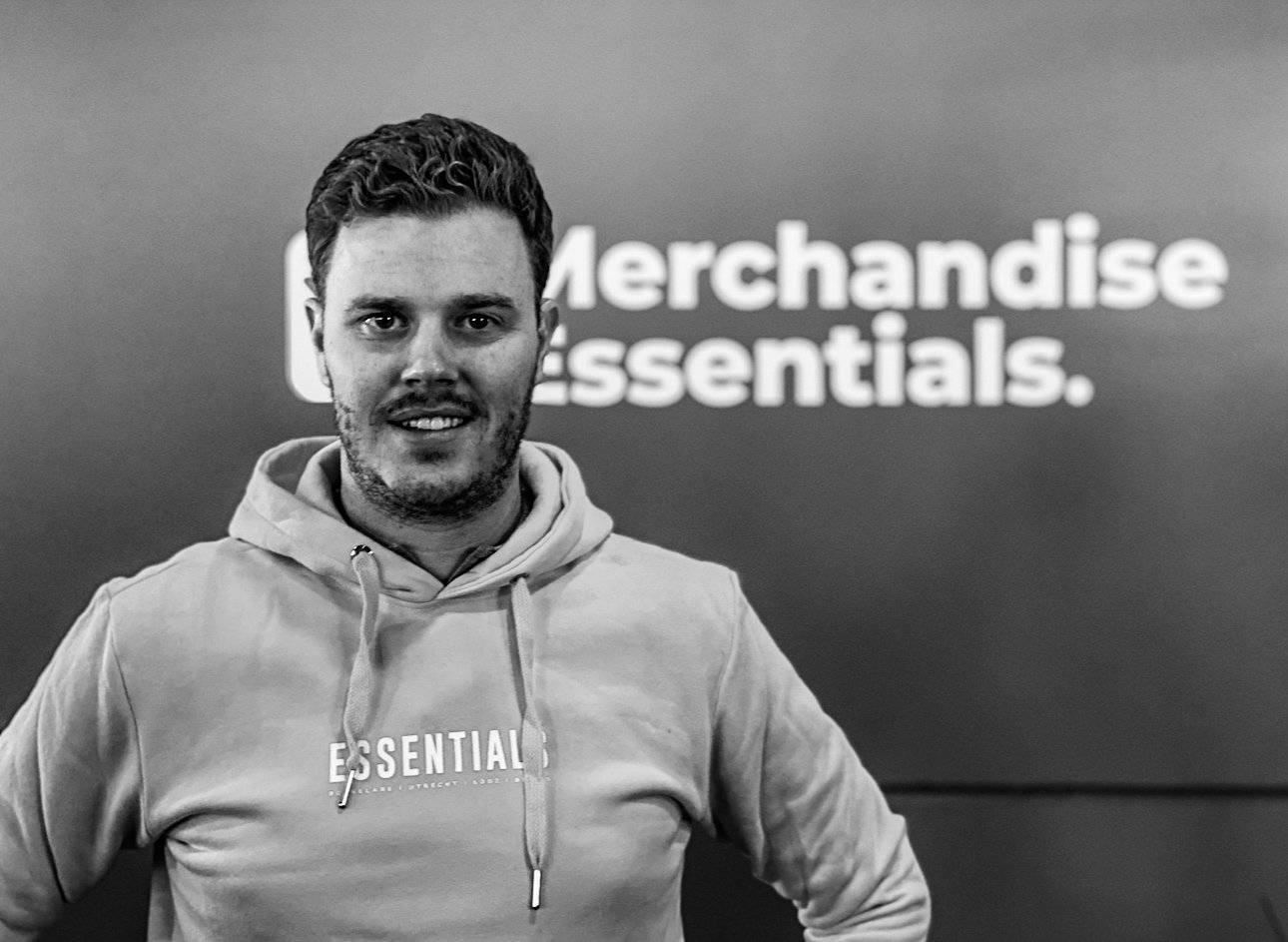 A man with short dark hair, facial hair, and wearing a hoodie with the word 'ESSENTIALS' on it, standing in front of a blurred wall with the words 'Merchandise essentials' partially visible.
