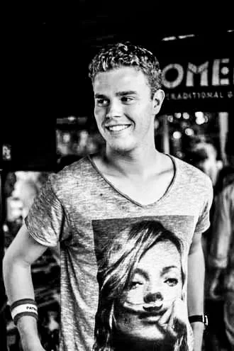 A smiling young man with curly hair wearing a graphic t-shirt with a woman's face on it, standing in a busy indoor setting.