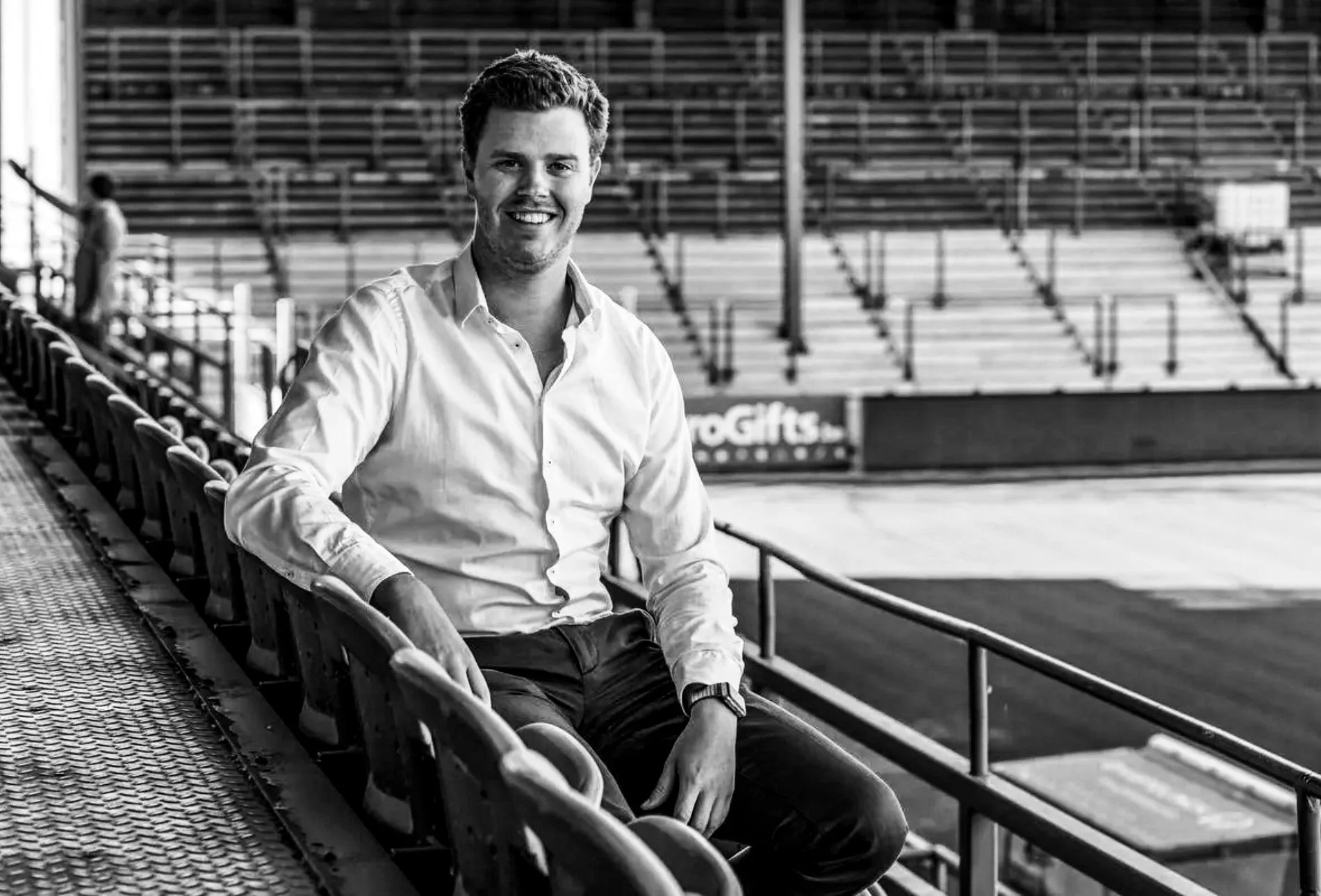 A smiling man in a white shirt sitting on stadium bleachers.