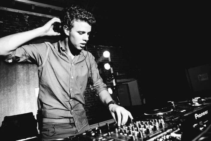 A young man DJ wearing headphones, standing behind a DJ mixer and turntables in a dark, brick-walled venue.