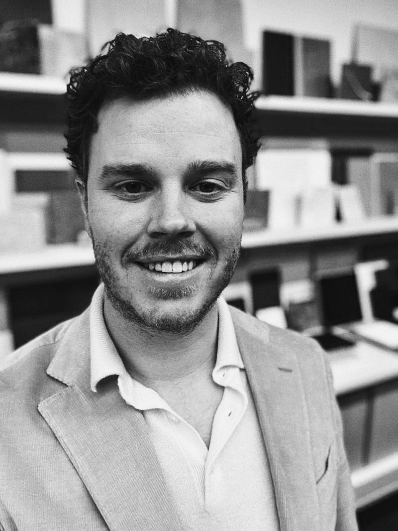 A smiling man with curly hair and beard, wearing a light-colored blazer and a collared shirt, standing in a store with shelves of books and electronics in the background.