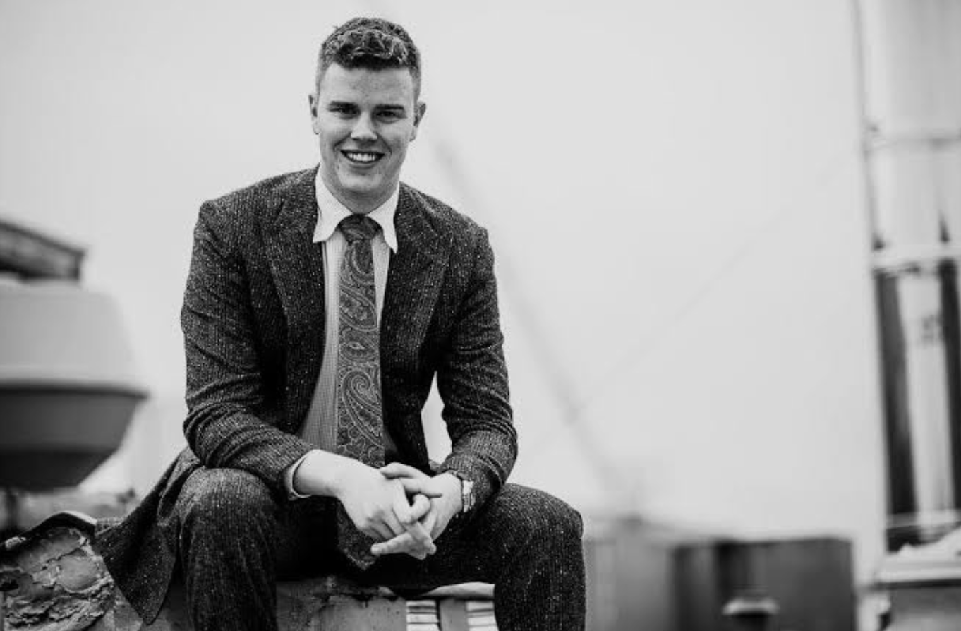 A young man in a suit and tie sits on a bench, smiling, in a black-and-white photo.