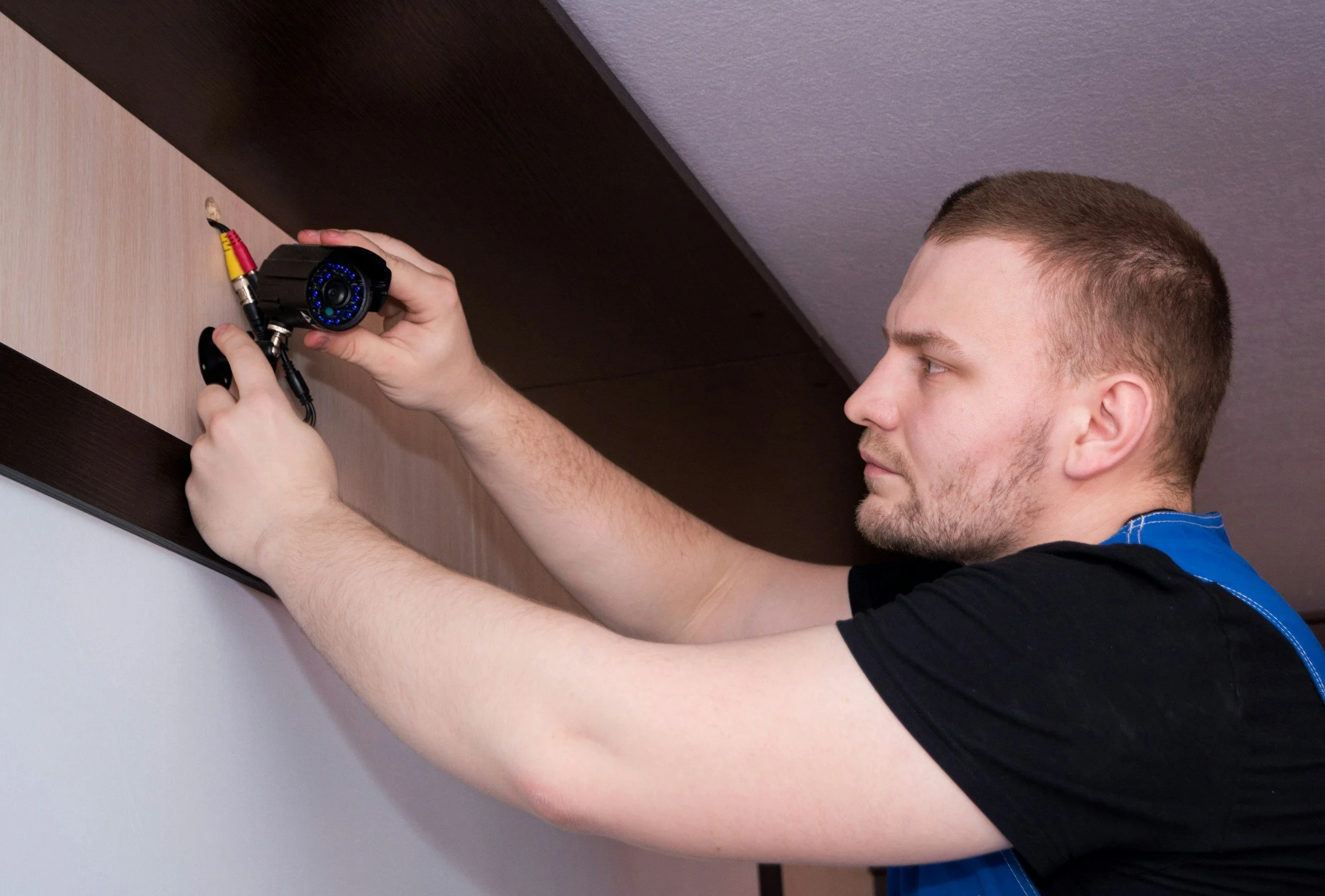 A man installing a security camera on a wall near the ceiling.