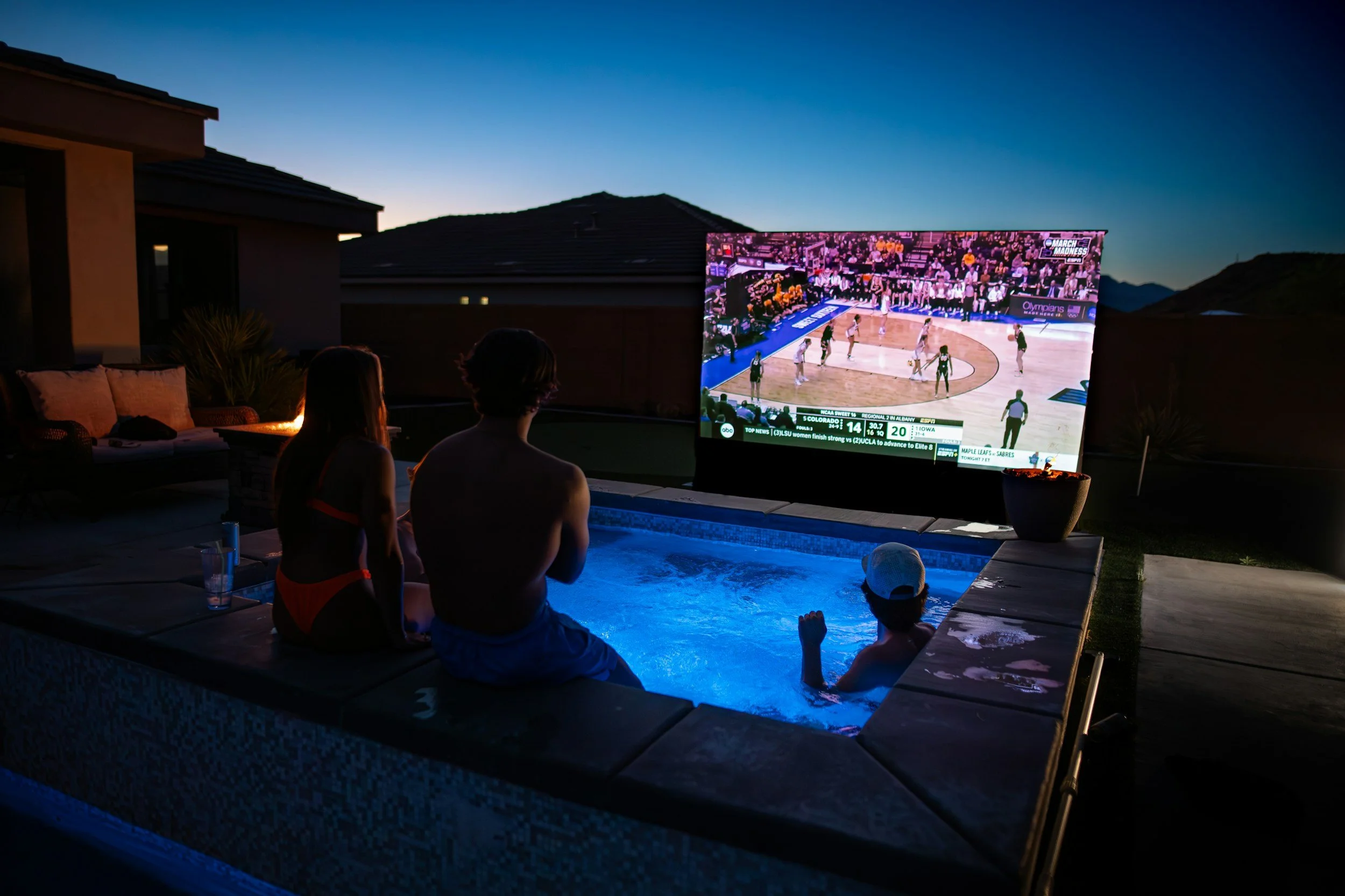 Three people sitting by a small pool, watching a basketball game on a large outdoor screen at dusk or night, with a house and mountains in the background.