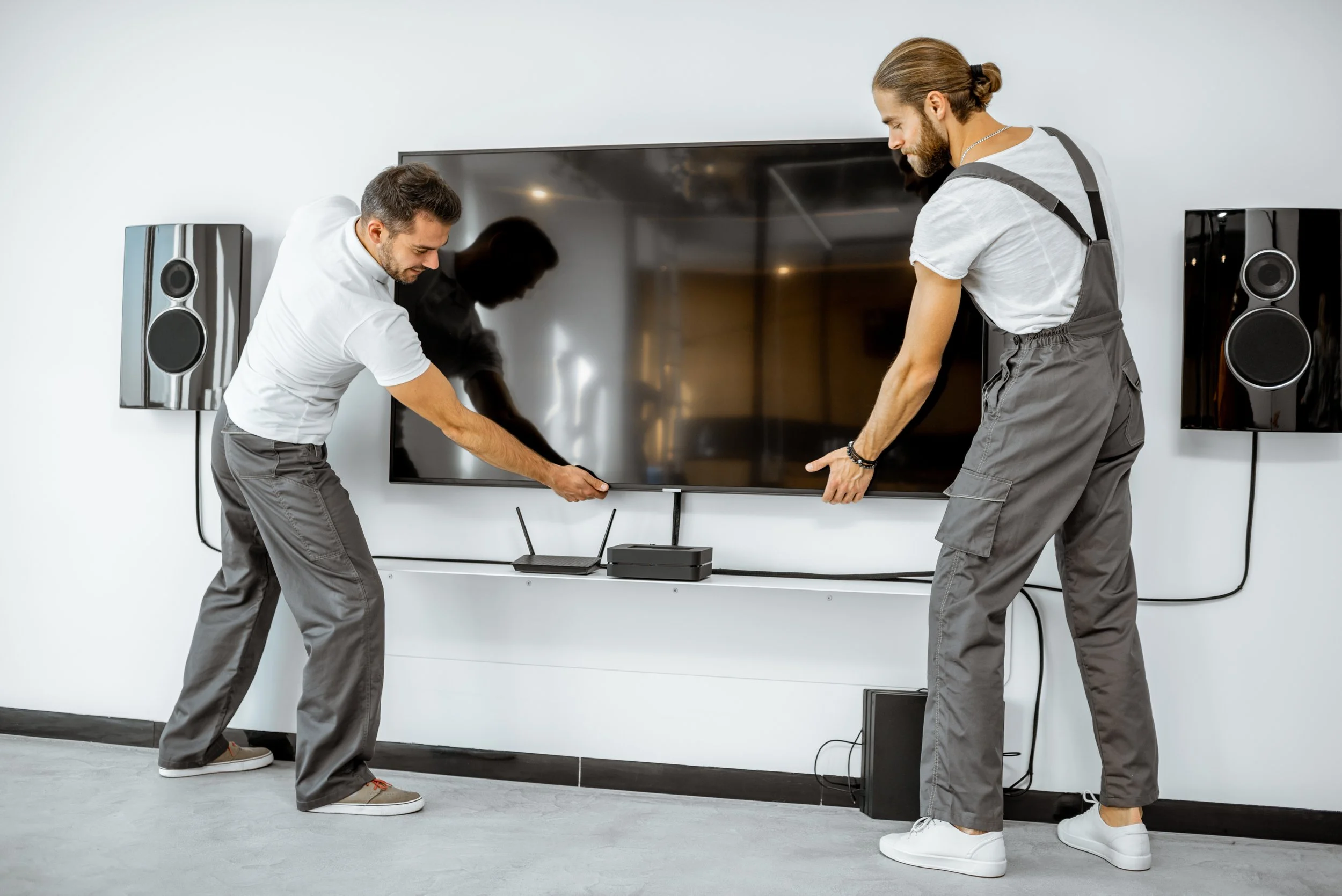 Two men are setting up or fixing a large flat-screen TV on a wall in a modern room, with speakers on either side and a TV box or router on a white shelf below.