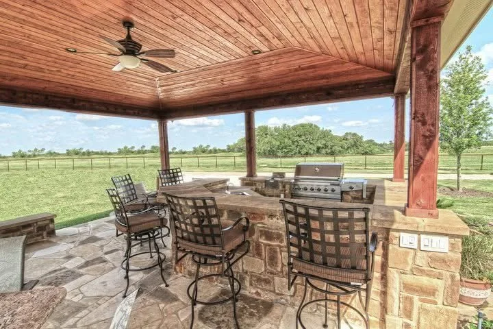 Outdoor covered patio with a brick counter, barstools, a grill, ceiling fan, and a view of a grassy field with trees in the background.