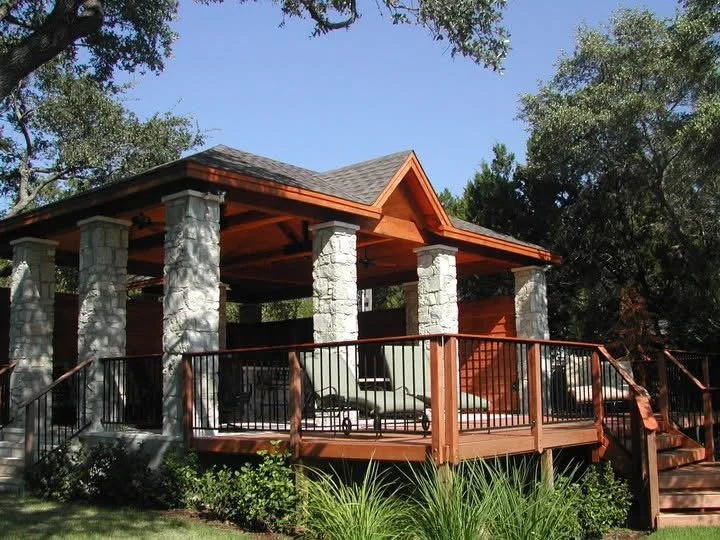 A covered outdoor patio with stone columns and wooden railings, surrounded by trees and greenery, under a clear blue sky.