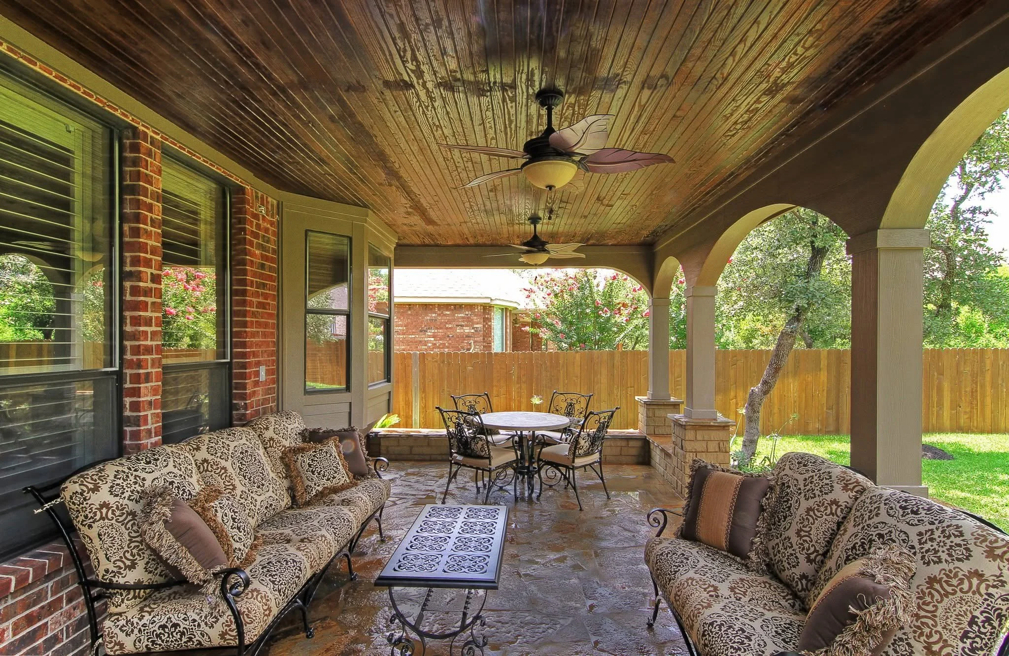 Covered patio with brick walls, a wooden ceiling with ceiling fans, a sofa set with patterned cushions, a round dining table with metal chairs, a stone floor, and a wooden fence with trees in the background.