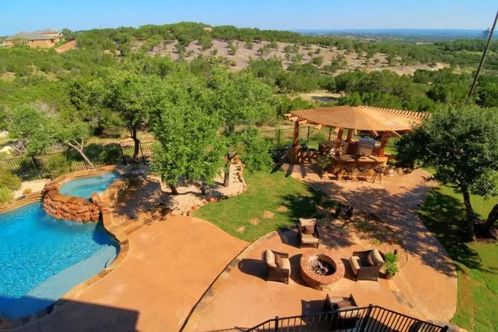 A backyard with a swimming pool, a fire pit surrounded by chairs, a shaded picnic area, and trees on a hilly landscape in the background.