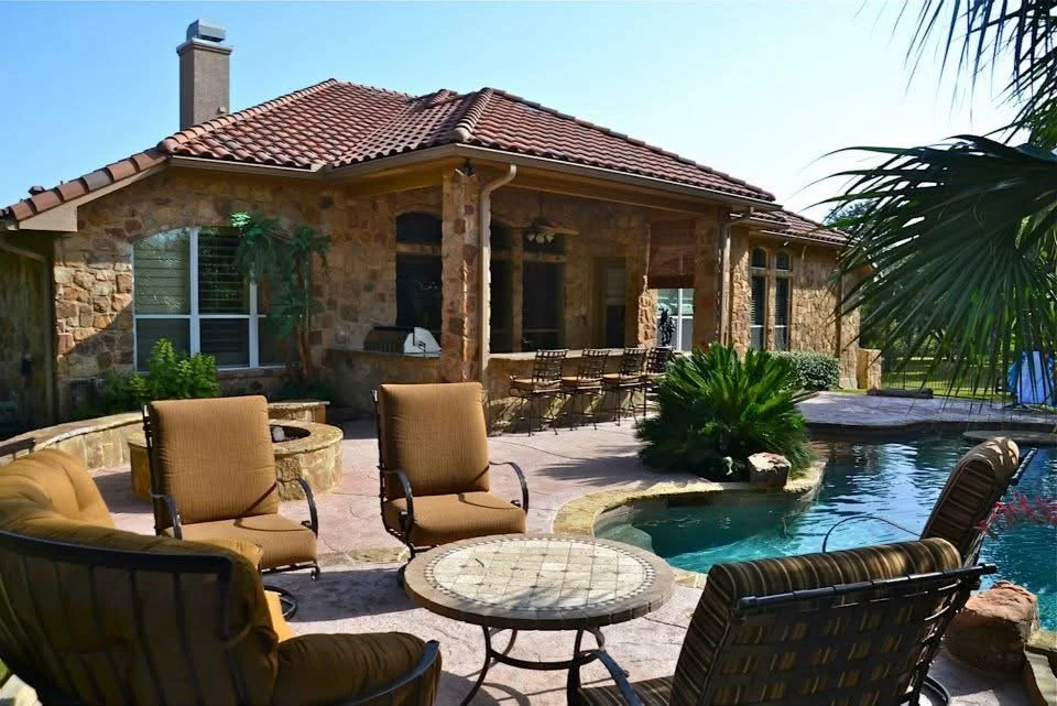Backyard patio with swimming pool, outdoor seating area with chairs and tables, stone house in the background, and lush greenery.