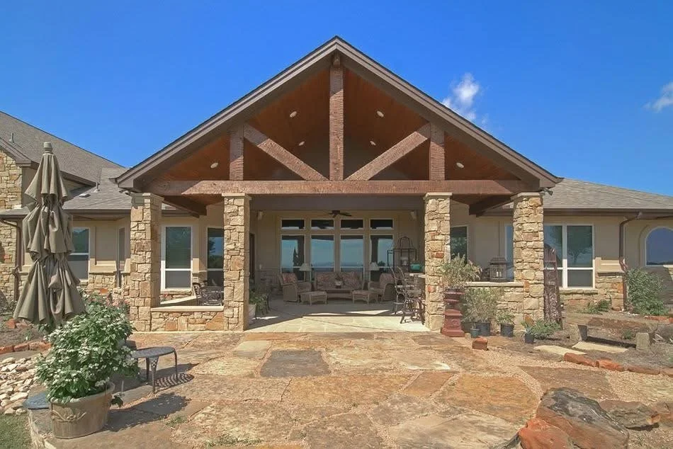 Backyard patio with stone and brick columns, seating area, large windows, and covered gable roof, surrounded by potted plants and outdoor furniture under a clear blue sky.